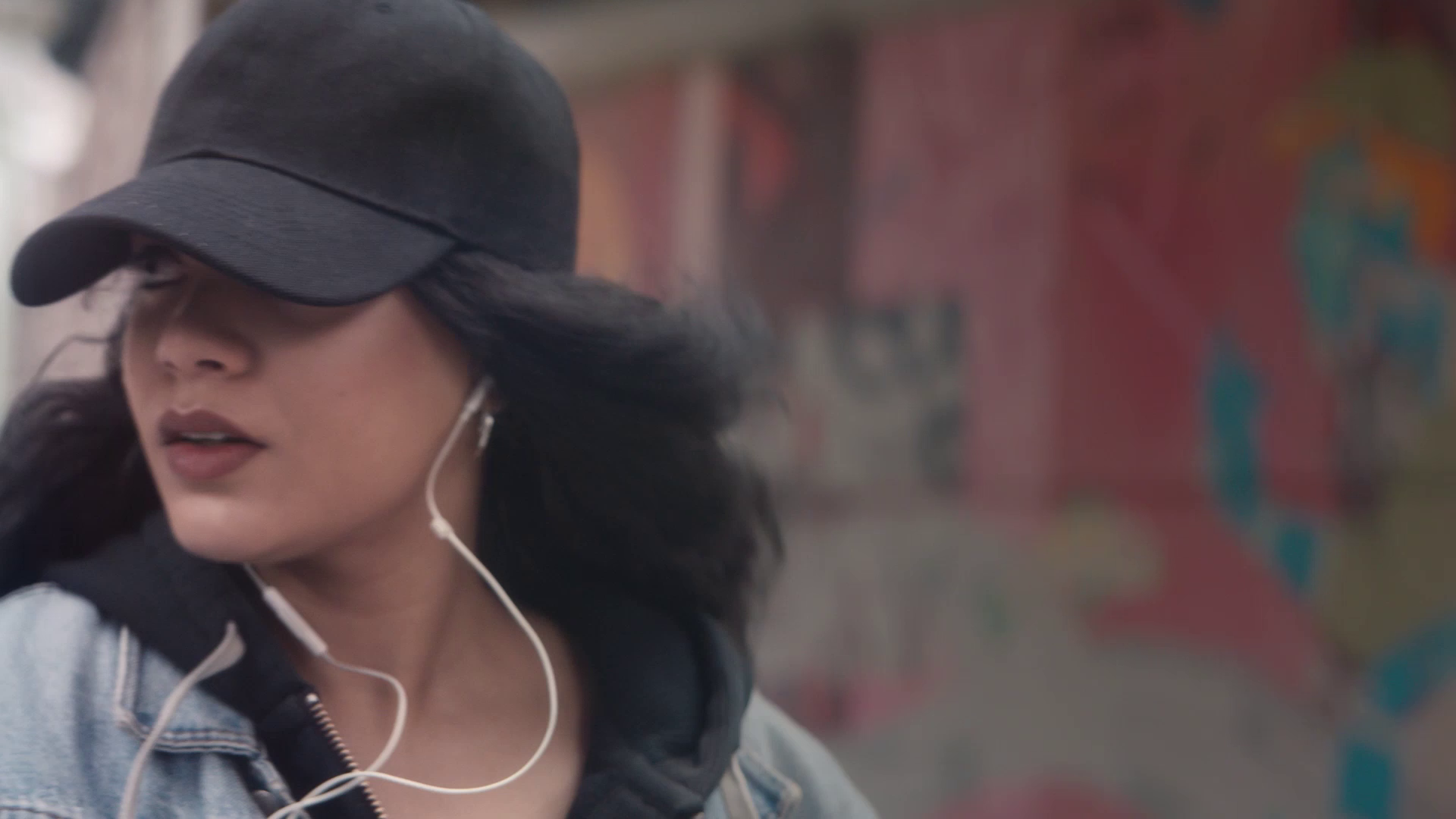 Woman wearing a black cap and denim jacket listening to music with white earphones.