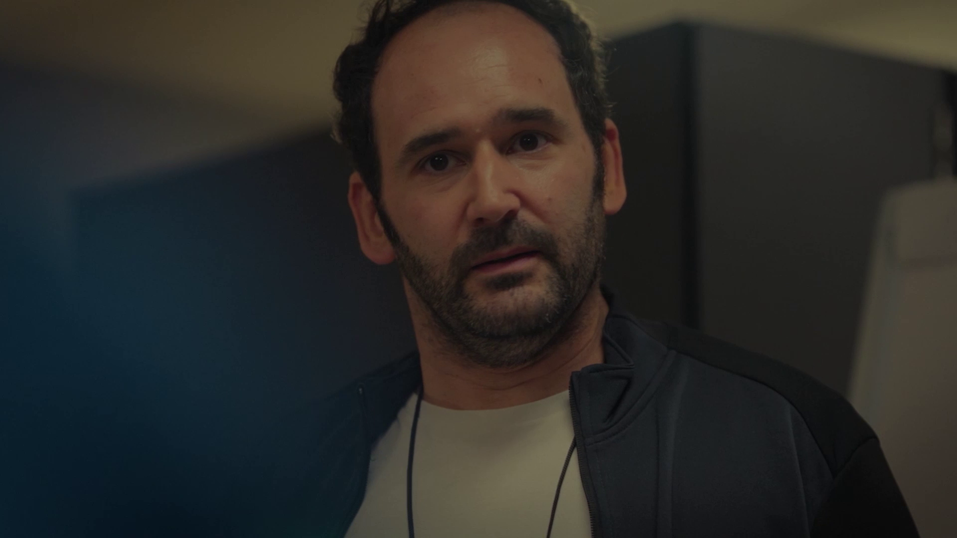 Close-up of a man with short curly hair and a beard wearing a black jacket over a white shirt indoors.