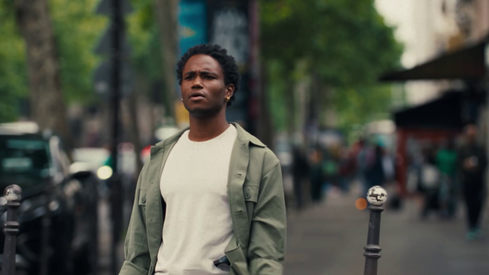 Young man wearing a green jacket and white shirt walking on a city sidewalk with blurred background.
