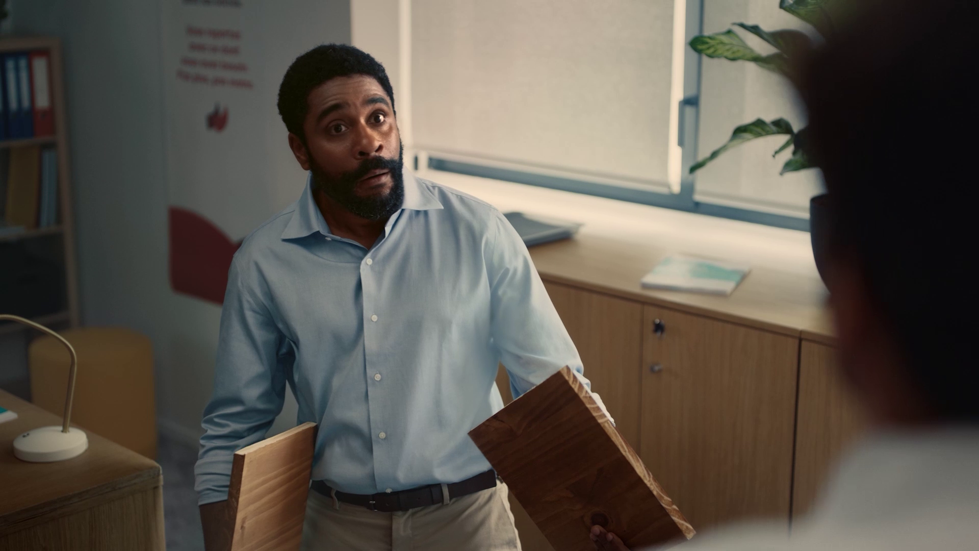 Man in light blue shirt holding two wooden planks with a surprised expression in an office.