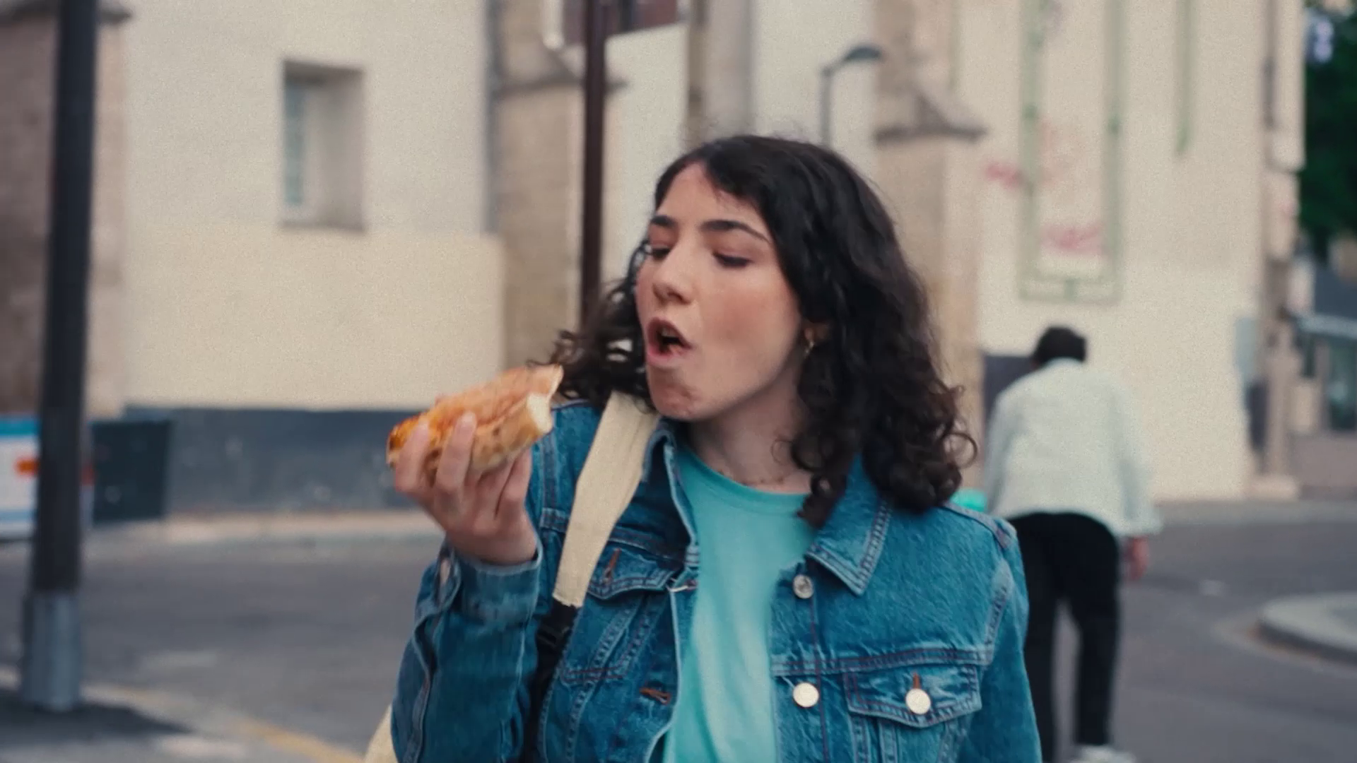 Young woman with curly hair wearing denim jacket eating a slice of pizza outdoors.