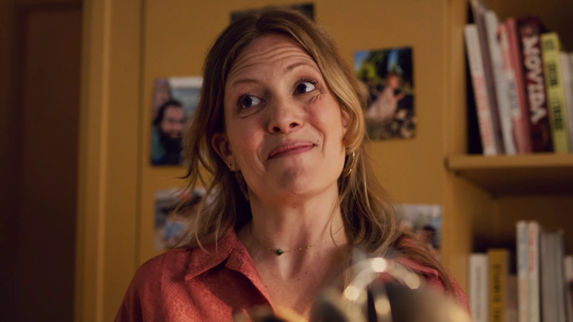 Woman with light brown hair and earrings wearing a patterned red shirt, smiling indoors with shelves and photos behind her.