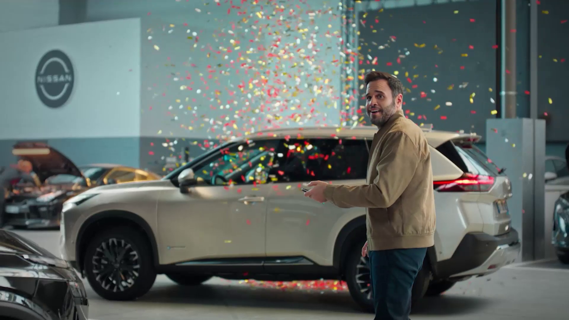 Smiling man holding a key fob near a beige Nissan SUV inside a showroom with confetti falling.