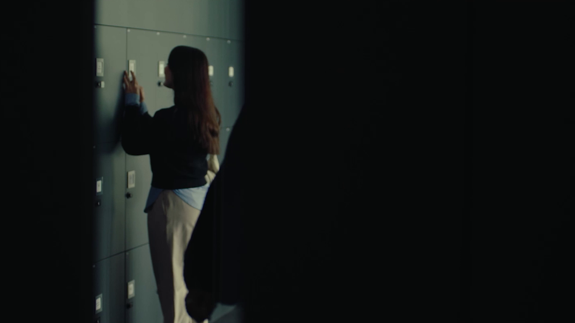Woman standing in front of lockers, interacting with one in a dimly lit hallway.