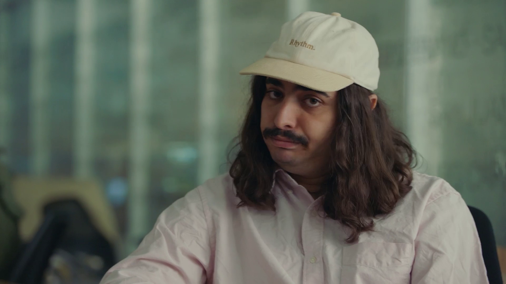 Man with long hair and mustache wearing a beige cap and light pink shirt, sitting indoors.