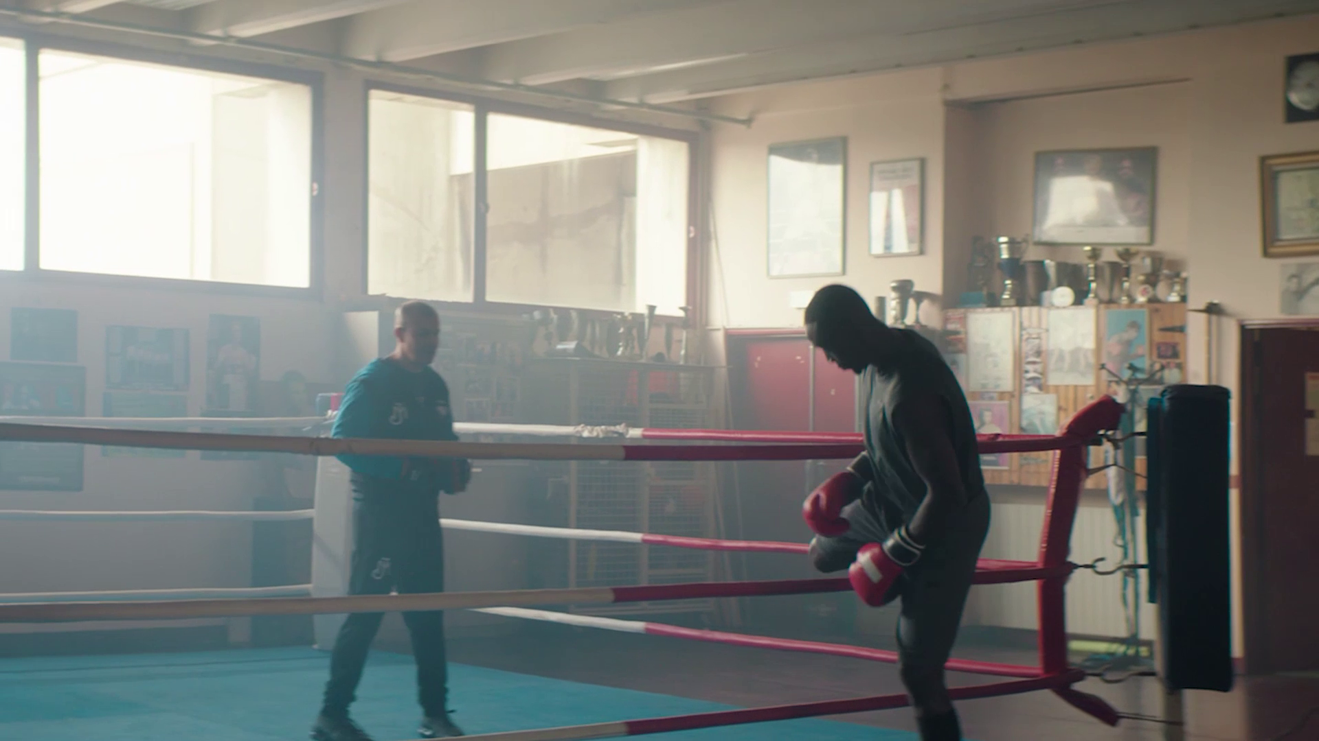 Boxer wearing gloves practices inside a boxing ring with a trainer in a gym.