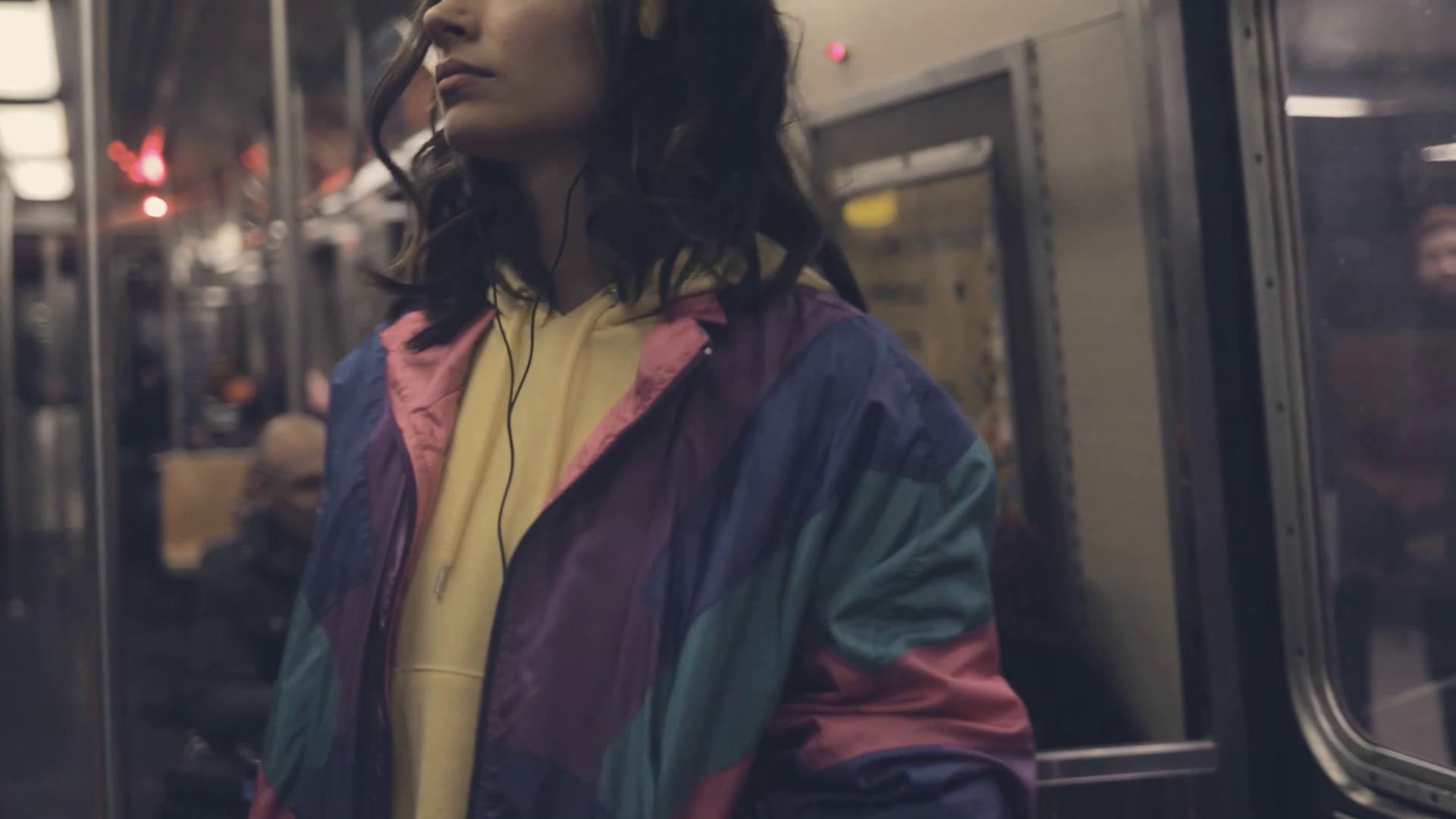 Woman with curly hair wearing headphones and a colorful jacket standing inside a subway train.
