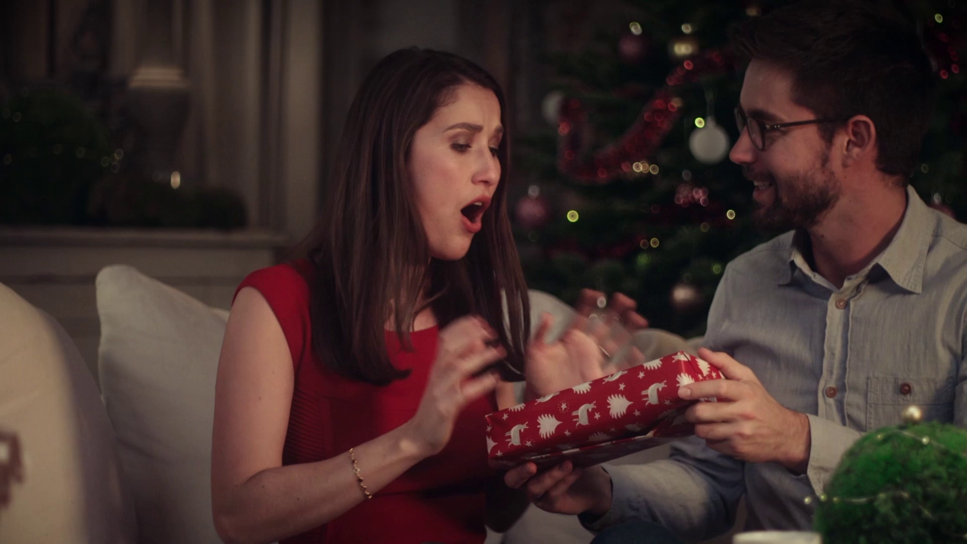 Woman in red dress surprised while receiving a Christmas gift from a man wearing glasses.