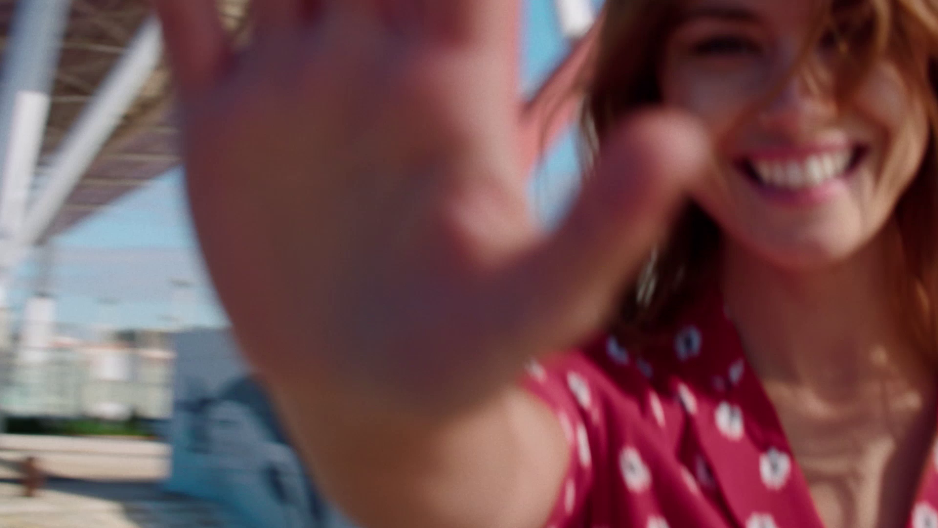 Woman smiling and reaching toward the camera outdoors on a sunny day.