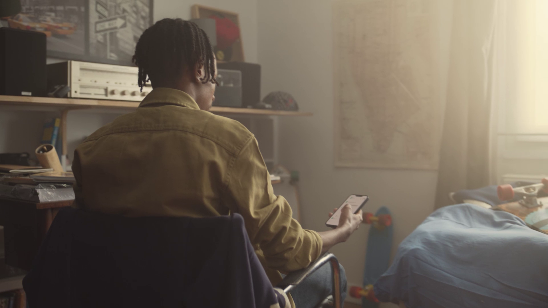 Person with braided hair sitting on a chair looking at a smartphone in a softly lit room with skateboard and map.