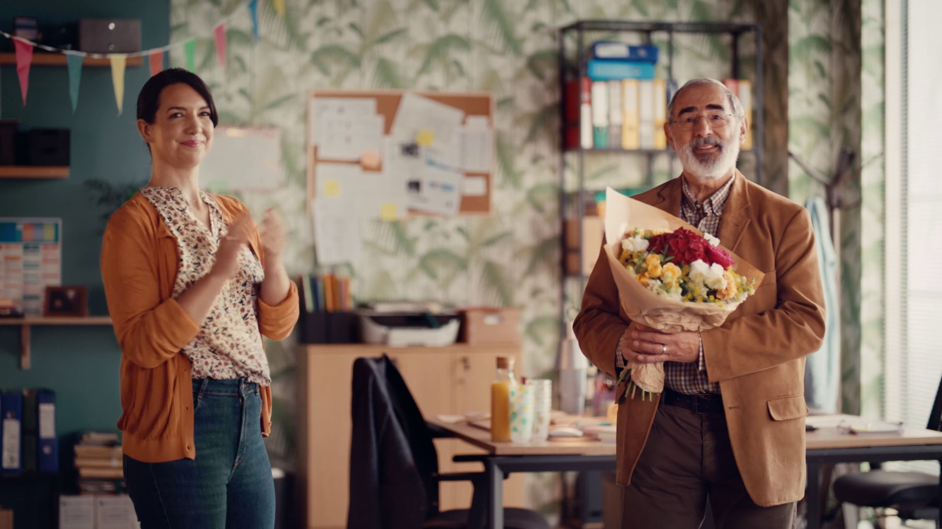 Older man holding a bouquet of flowers while a woman claps in an office setting.