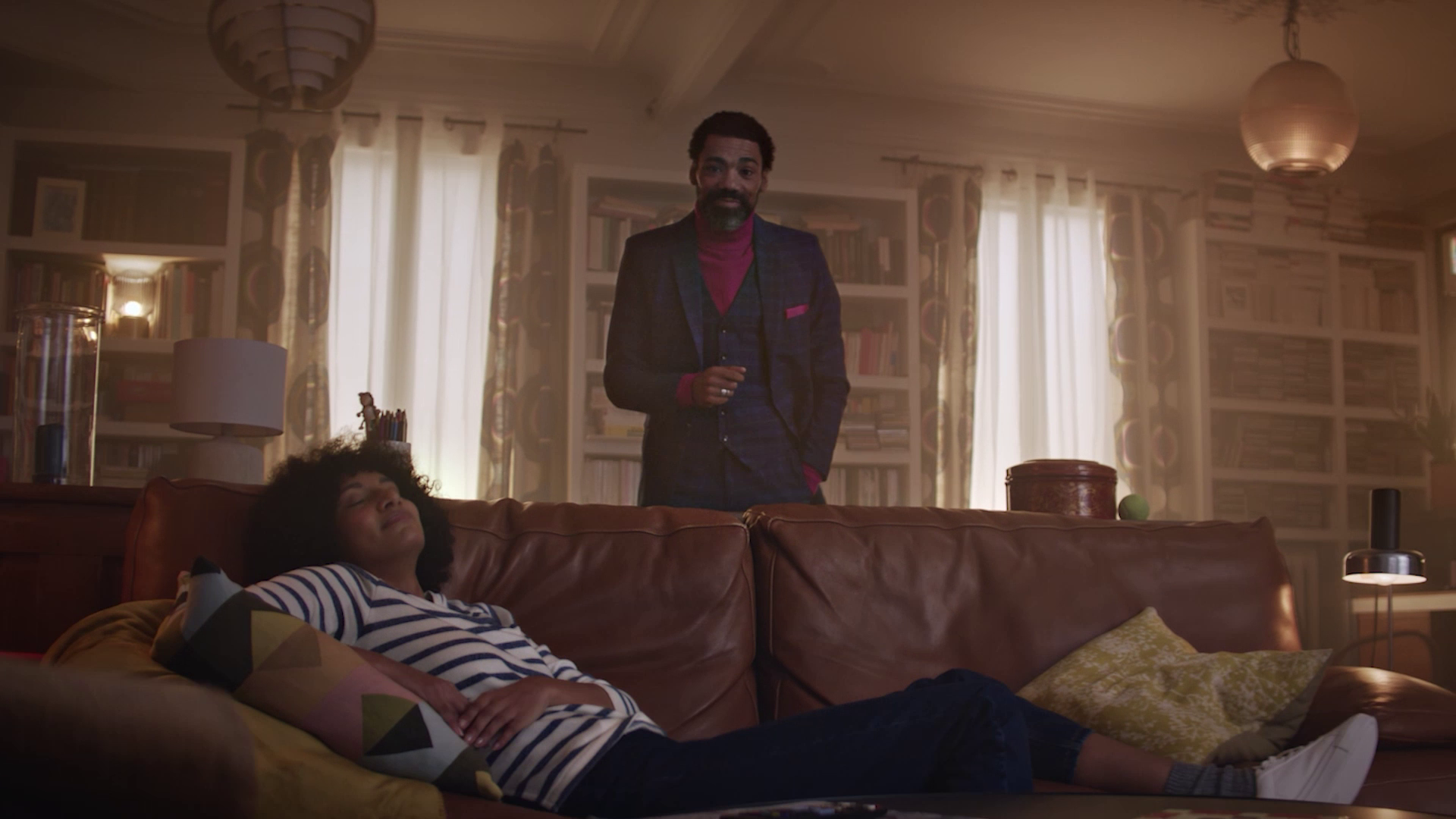 Woman relaxing on a brown leather couch with a man standing behind her in a cozy, book-filled living room.