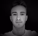 Black and white portrait of a young man with short hair looking straight at the camera against a black background.