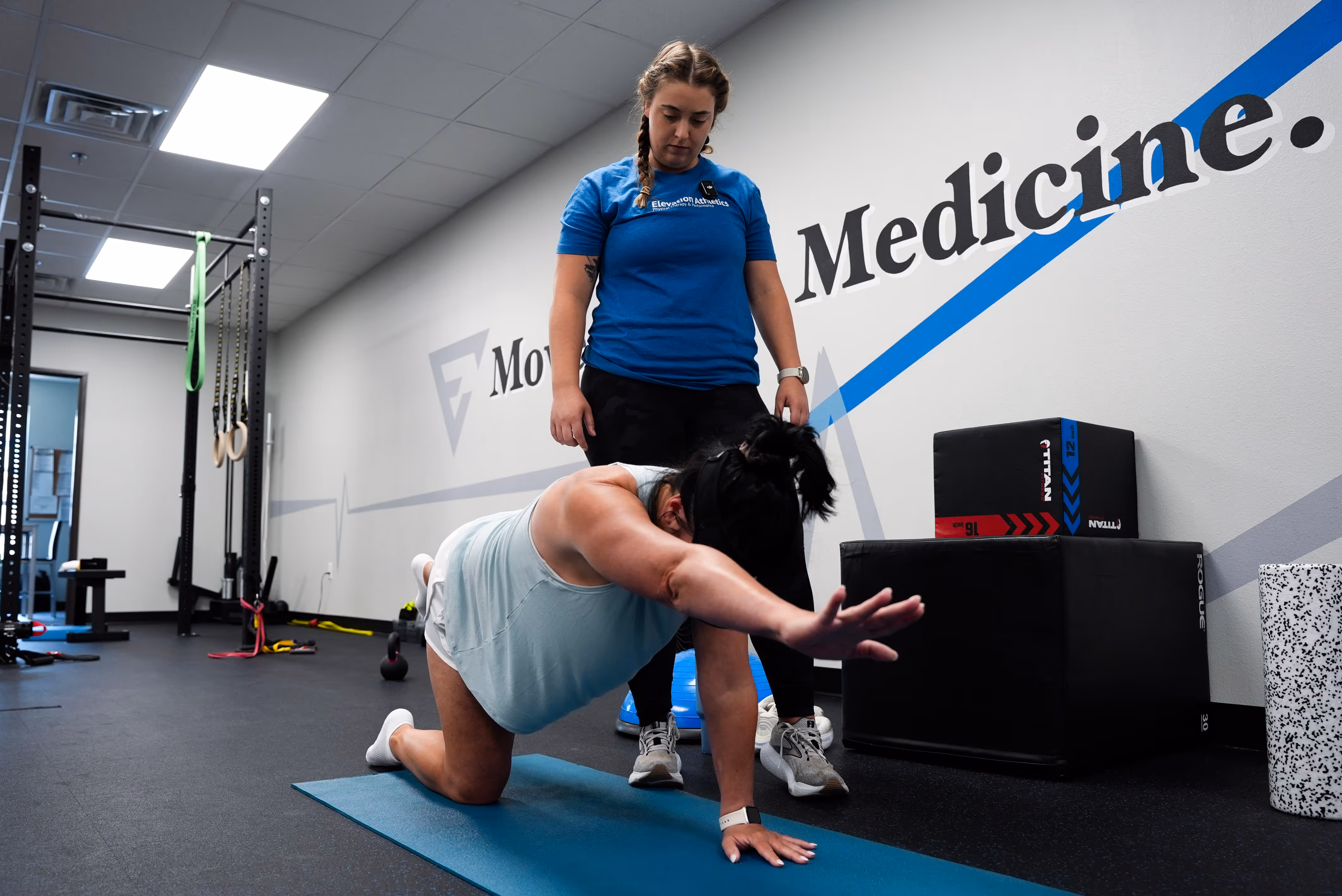 A woman on a blue exercise mat stretching one arm forward and opposite knee back while a trainer in a blue shirt stands behind her in a gym with exercise equipment and a wall graphic that says 'Medicine.' elevation-athletics-pt