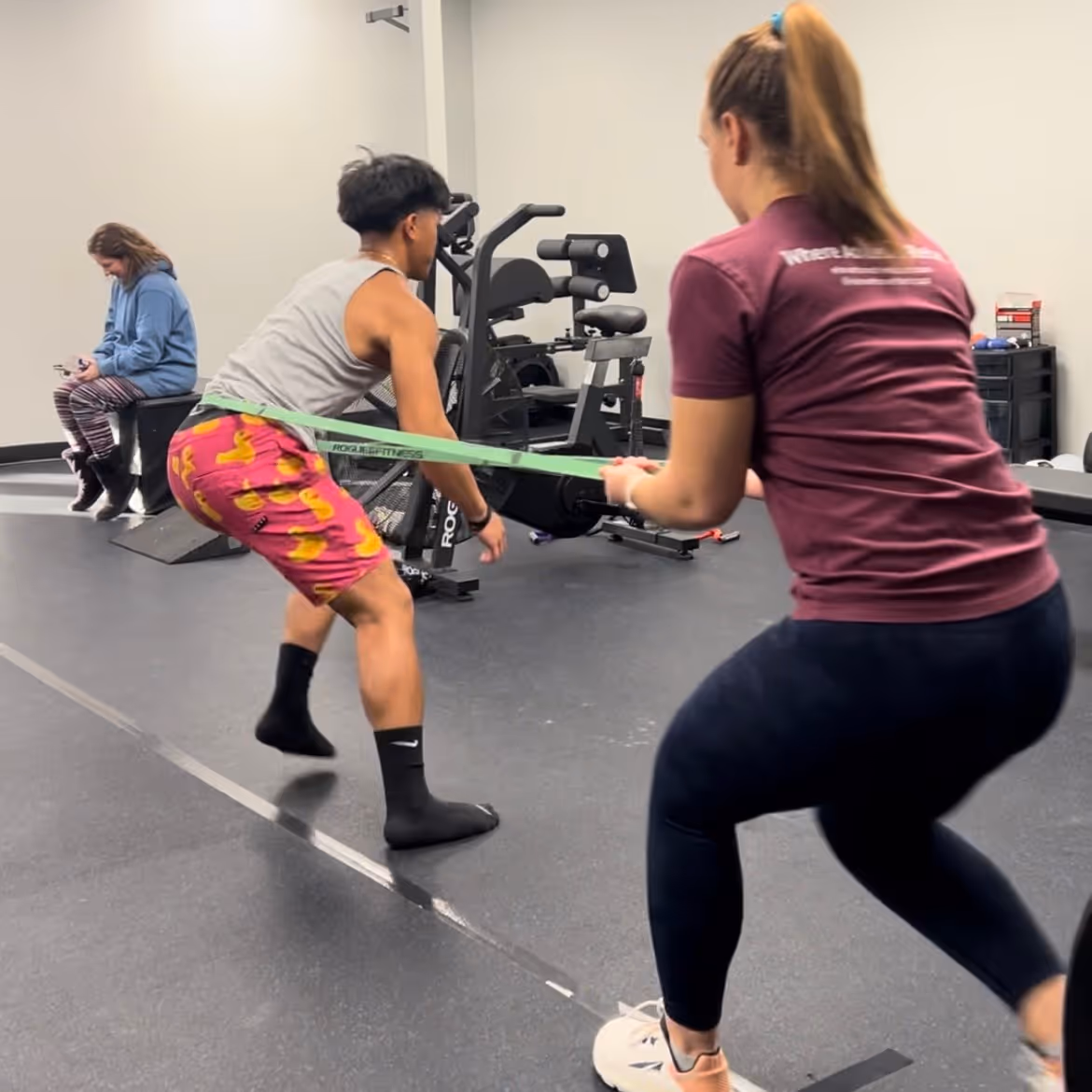 Two people exercising in a gym with a resistance band while a third person sits in the background. elevation-athletics-pt