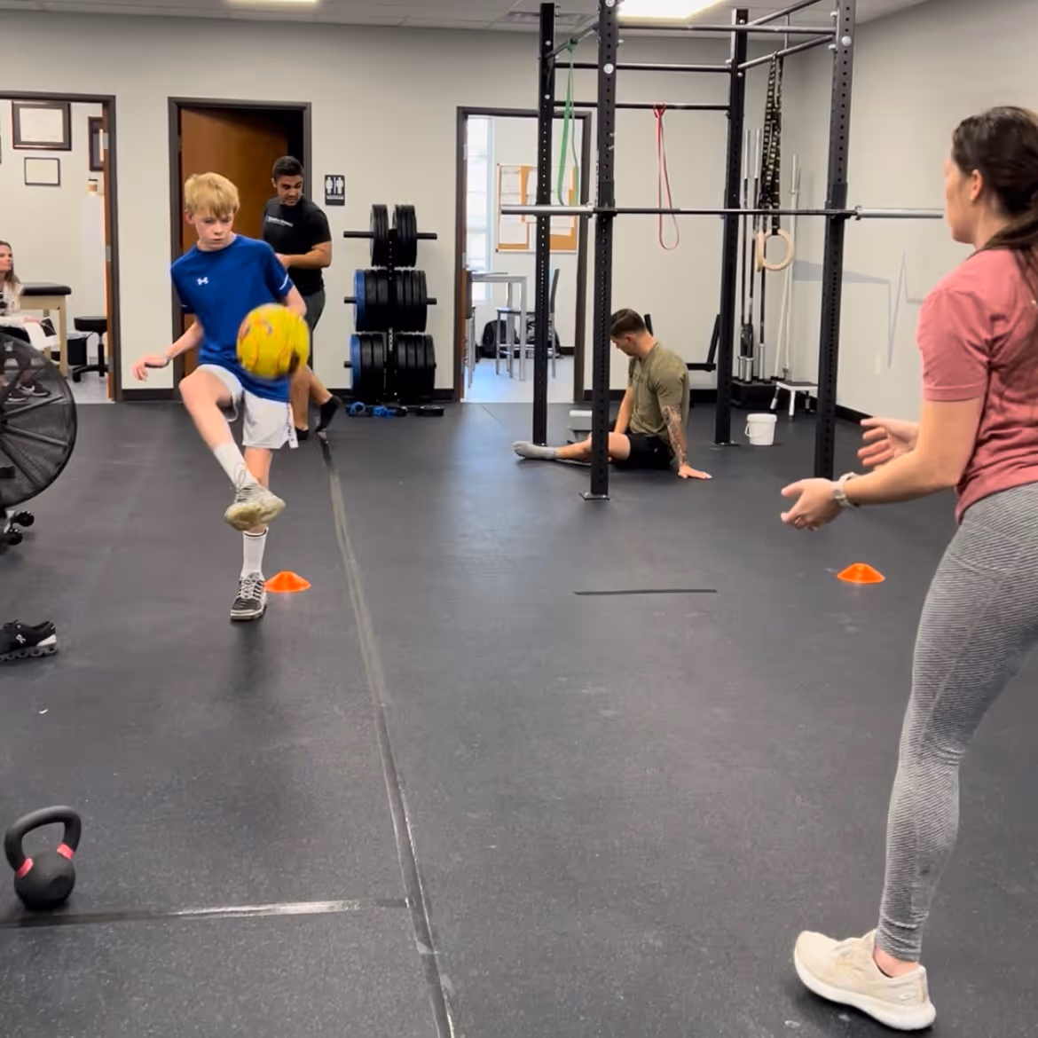 A boy in blue shirt kicking a yellow soccer ball indoors with a man and woman observing in a gym setting. elevation-athletics-pt