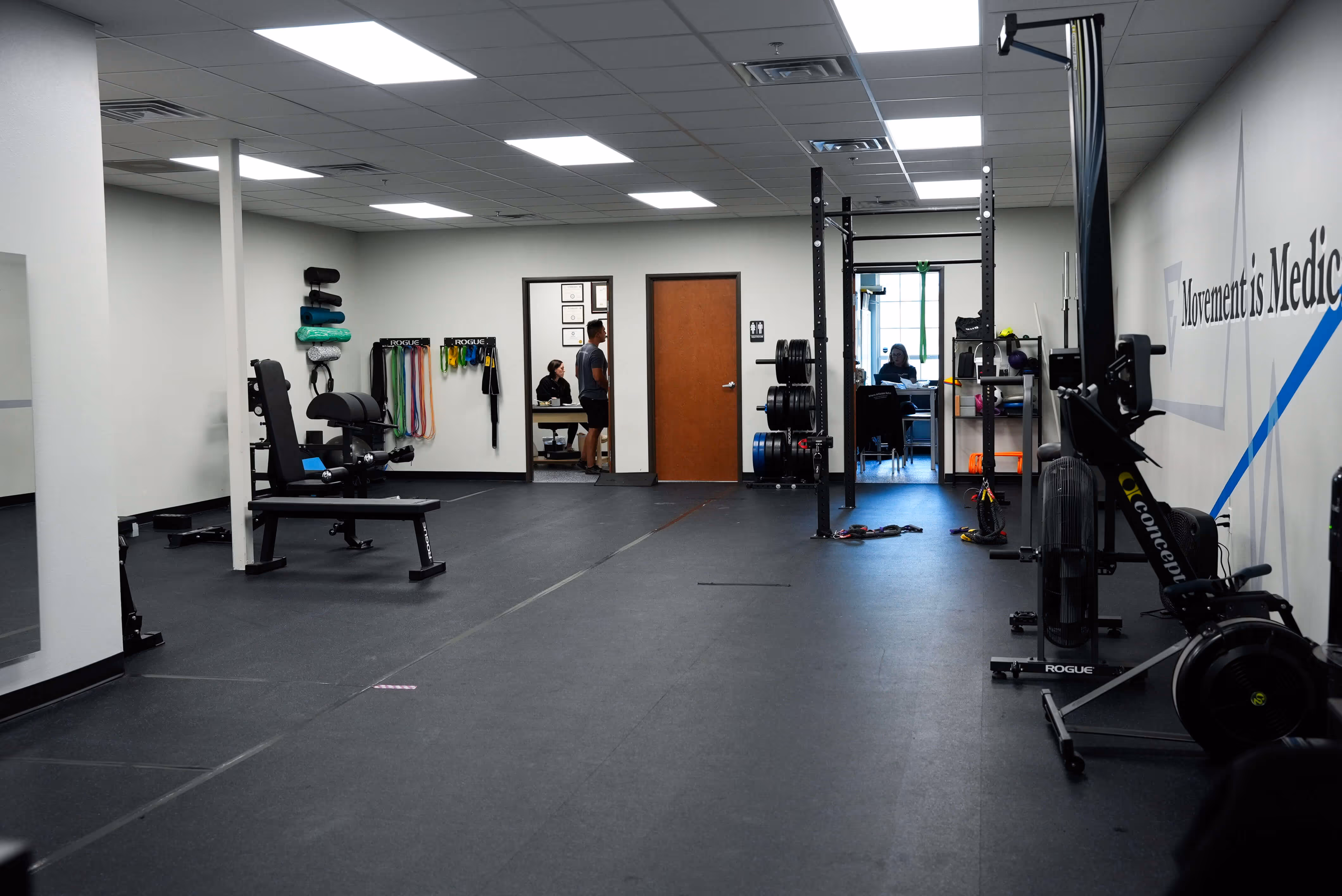 Modern gym room with black rubber flooring, weight bench, resistance bands, weight plates, and exercise machines, two people talking in an office in the background. elevation-athletics-pt
