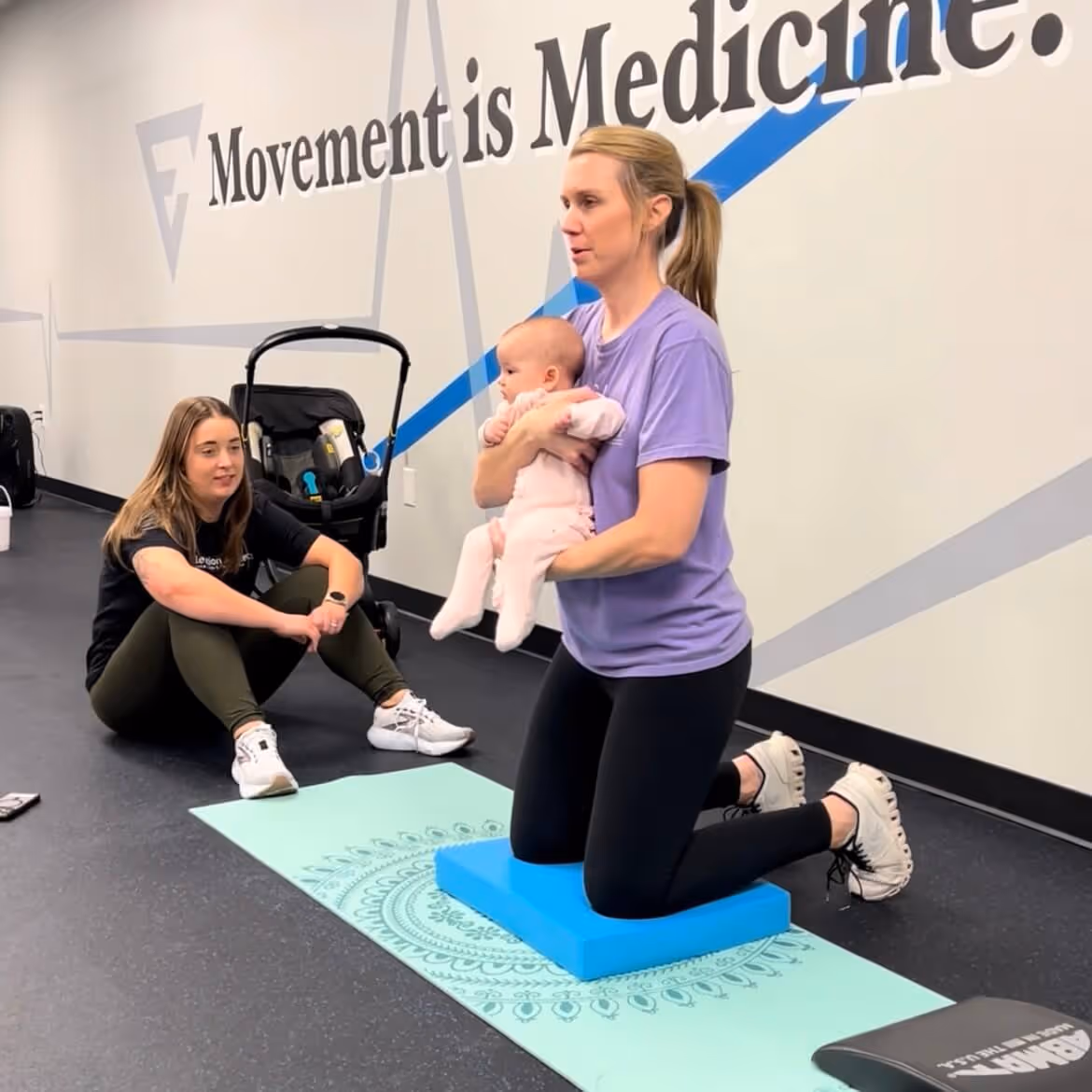 Woman kneeling on a blue foam pad holding a baby while another woman sits on the floor watching in a fitness studio. elevation-athletics-pt