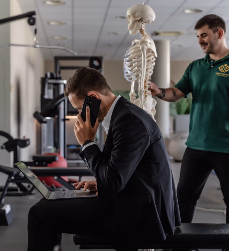 Ein Mann im Anzug telefoniert und arbeitet an einem Laptop, während ein Mann in grüner Arbeitskleidung neben einem menschlichen Skelett steht.