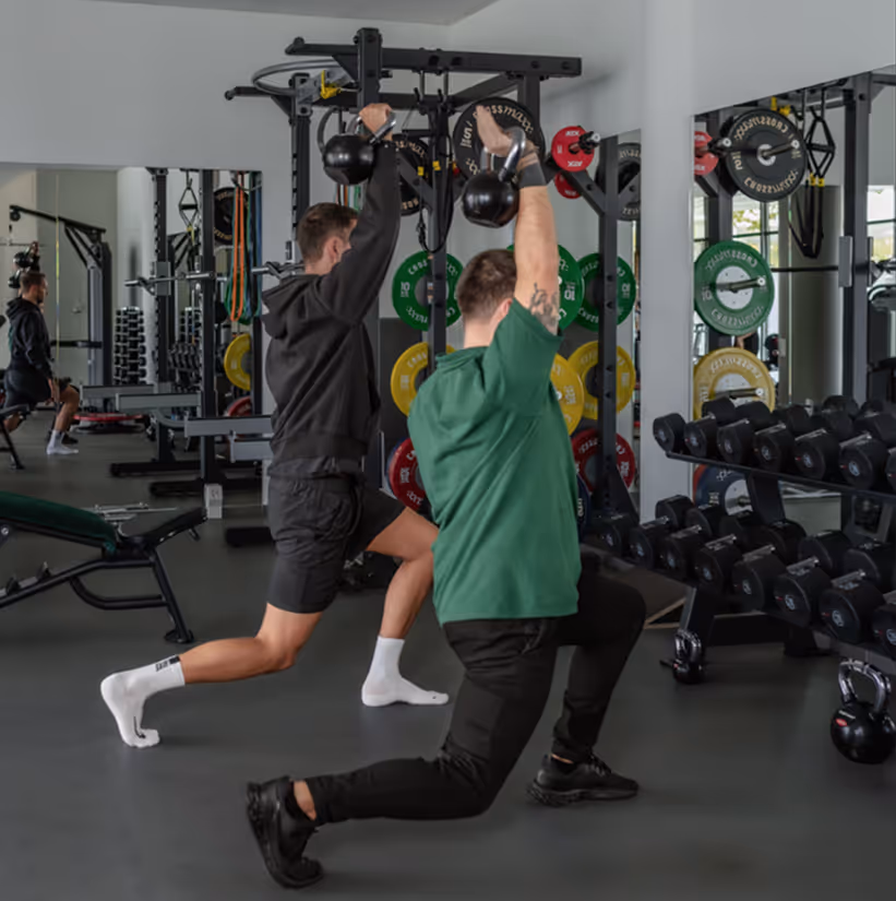 Zwei Männer machen Ausfallschritte mit Kettlebells über Kopf in einem Fitnessstudio.