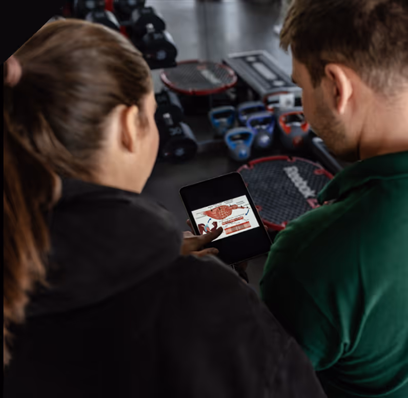 Zwei Personen betrachten eine Grafik eines Muskels auf einem Tablet in einem Fitnessstudio.