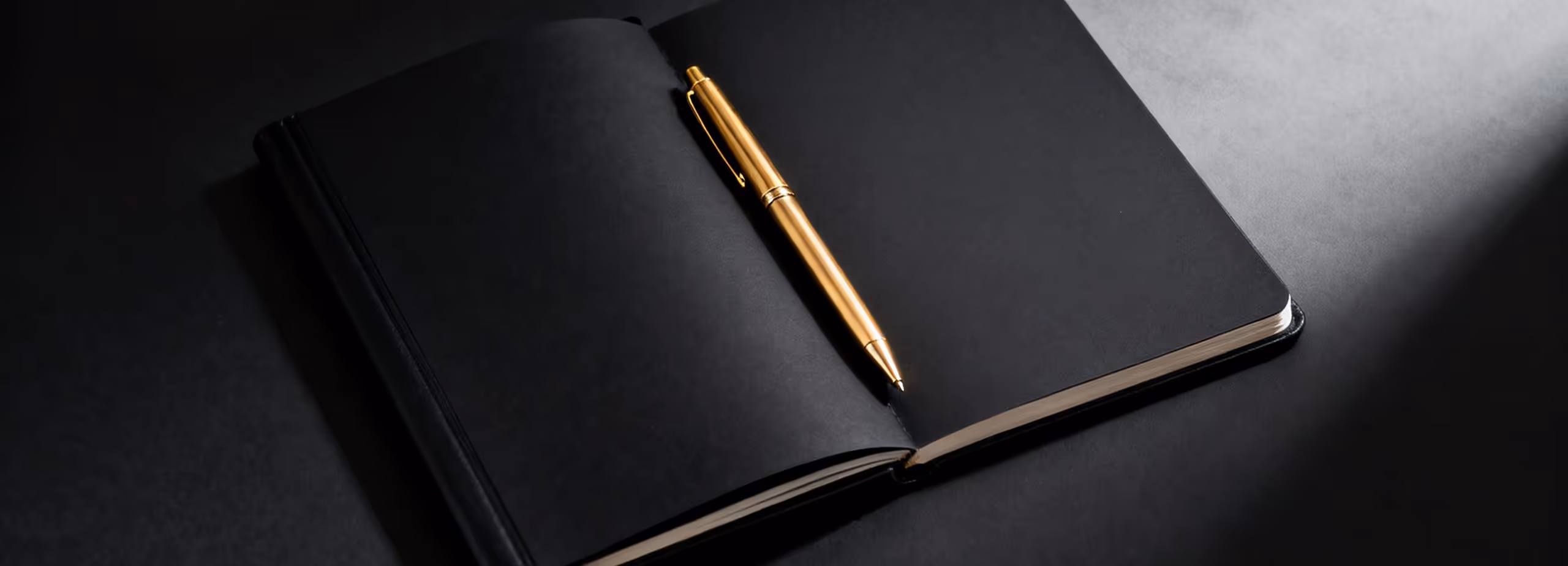 Ein aufgeschlagenes schwarzes Notizbuch mit einem goldenen Kugelschreiber darauf, auf schwarzem Untergrund mit Lichtreflexion.