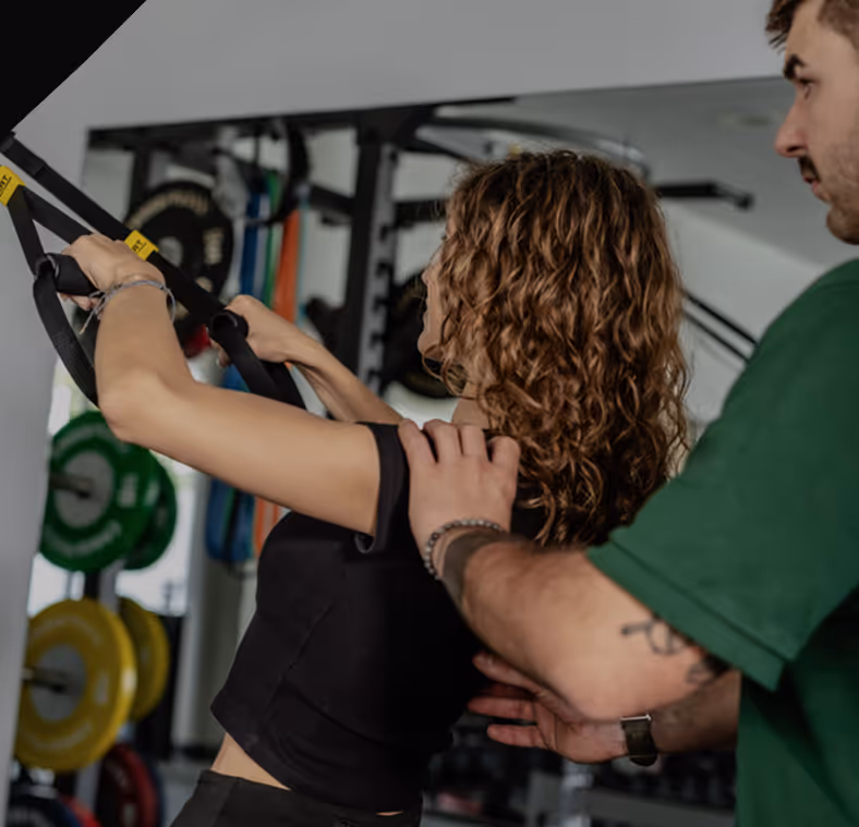 Frau trainiert mit Widerstandsbändern im Fitnessstudio, während ein Trainer sie an Schulter und Rücken unterstützt.