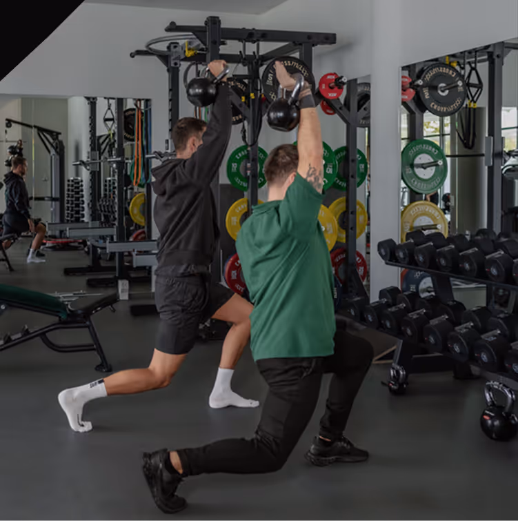 Zwei Männer machen Ausfallschritte und heben gleichzeitig Kettlebells über den Kopf in einem Fitnessstudio.