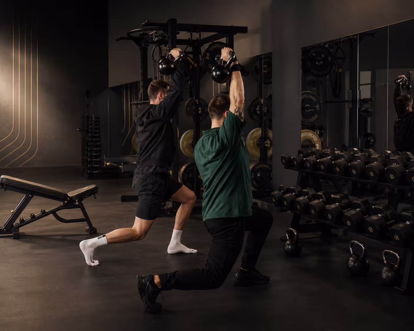 Zwei Männer machen Ausfallschritte mit Kettlebells in einem Fitnessstudio neben Spiegeln und Gewichten.