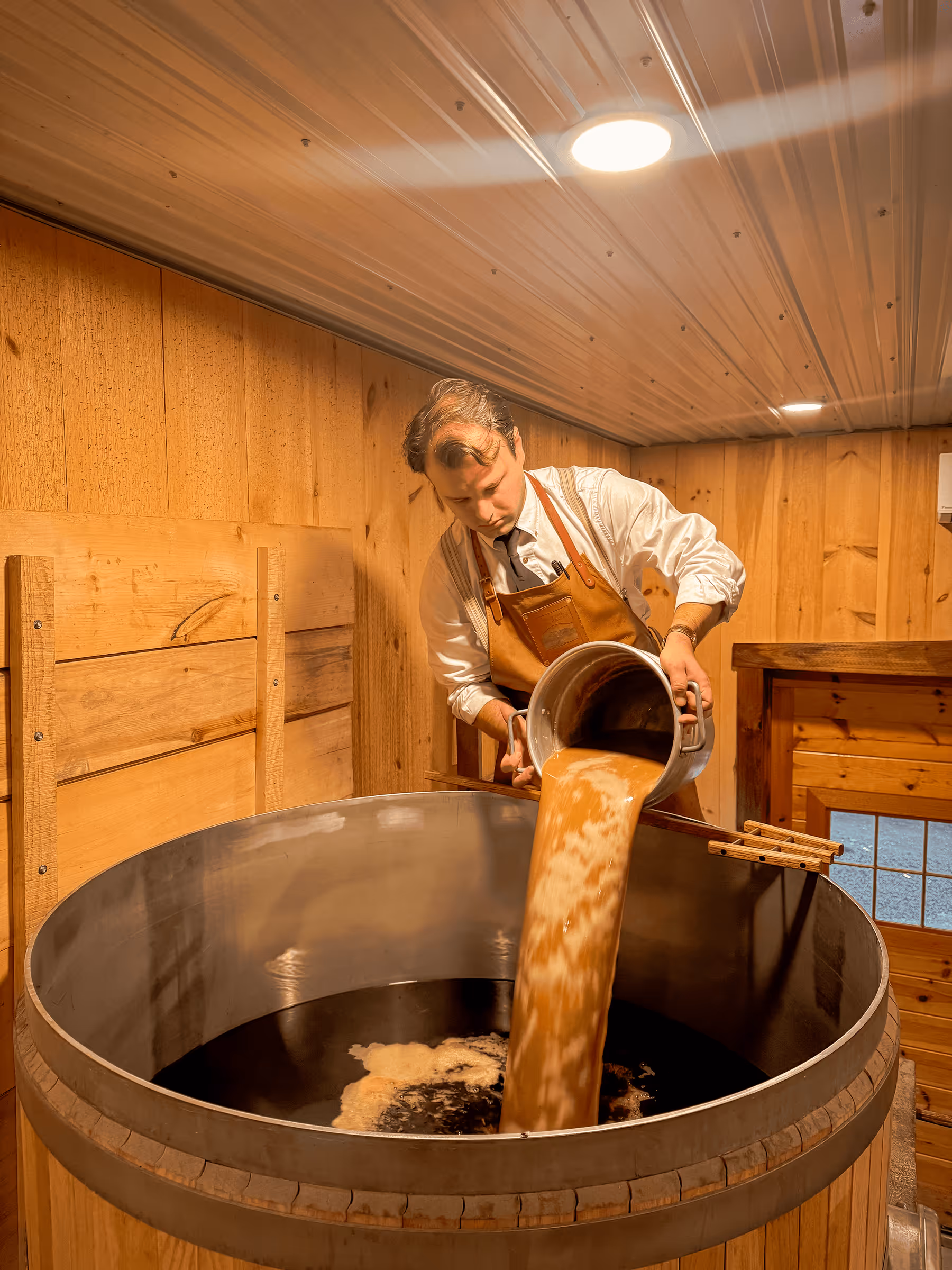 Craftsman in apron pouring liquid into large wooden brewing barrel