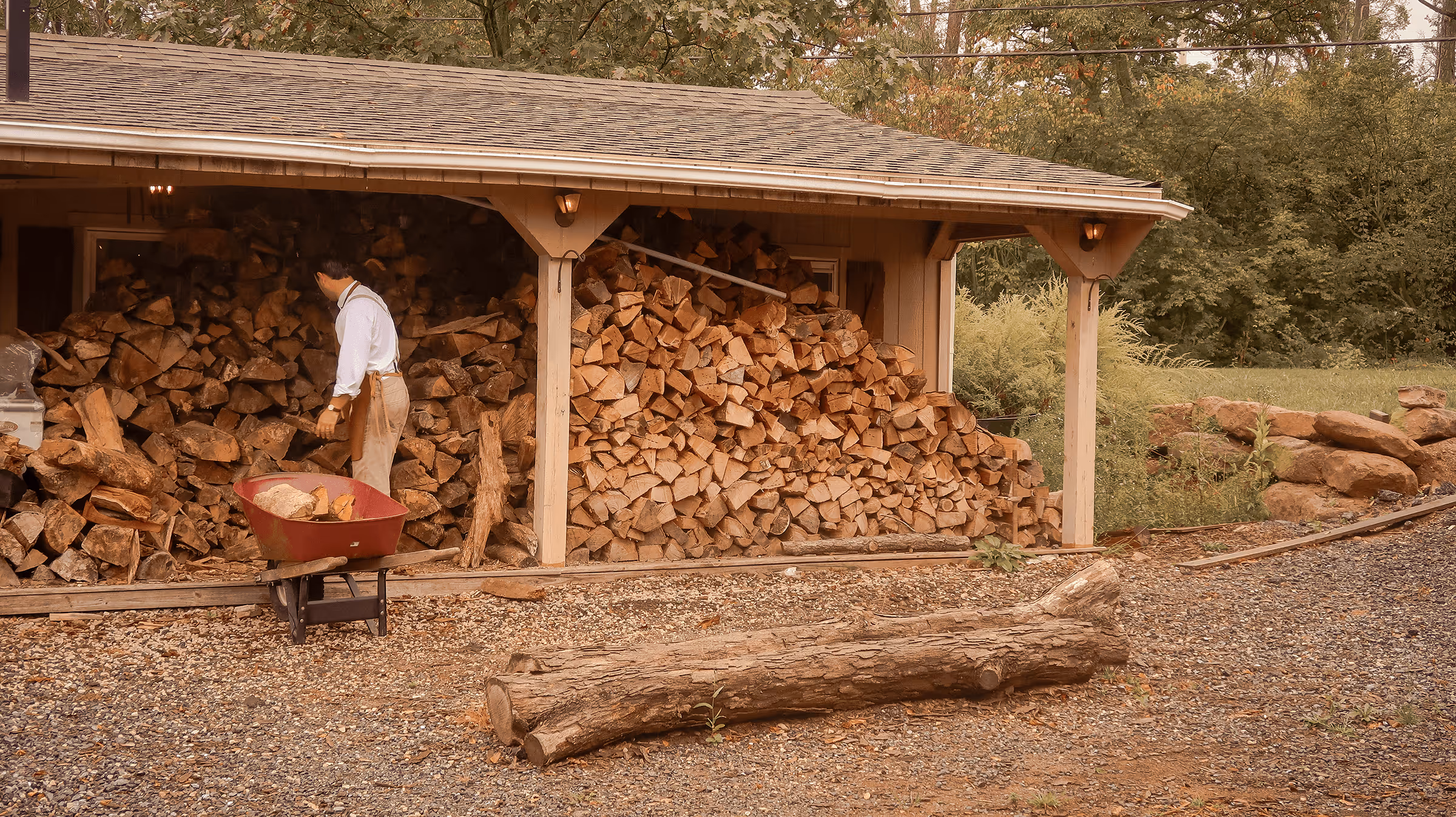 Woodshed with stacked firewood and person preparing wood in a red wheelbarrow