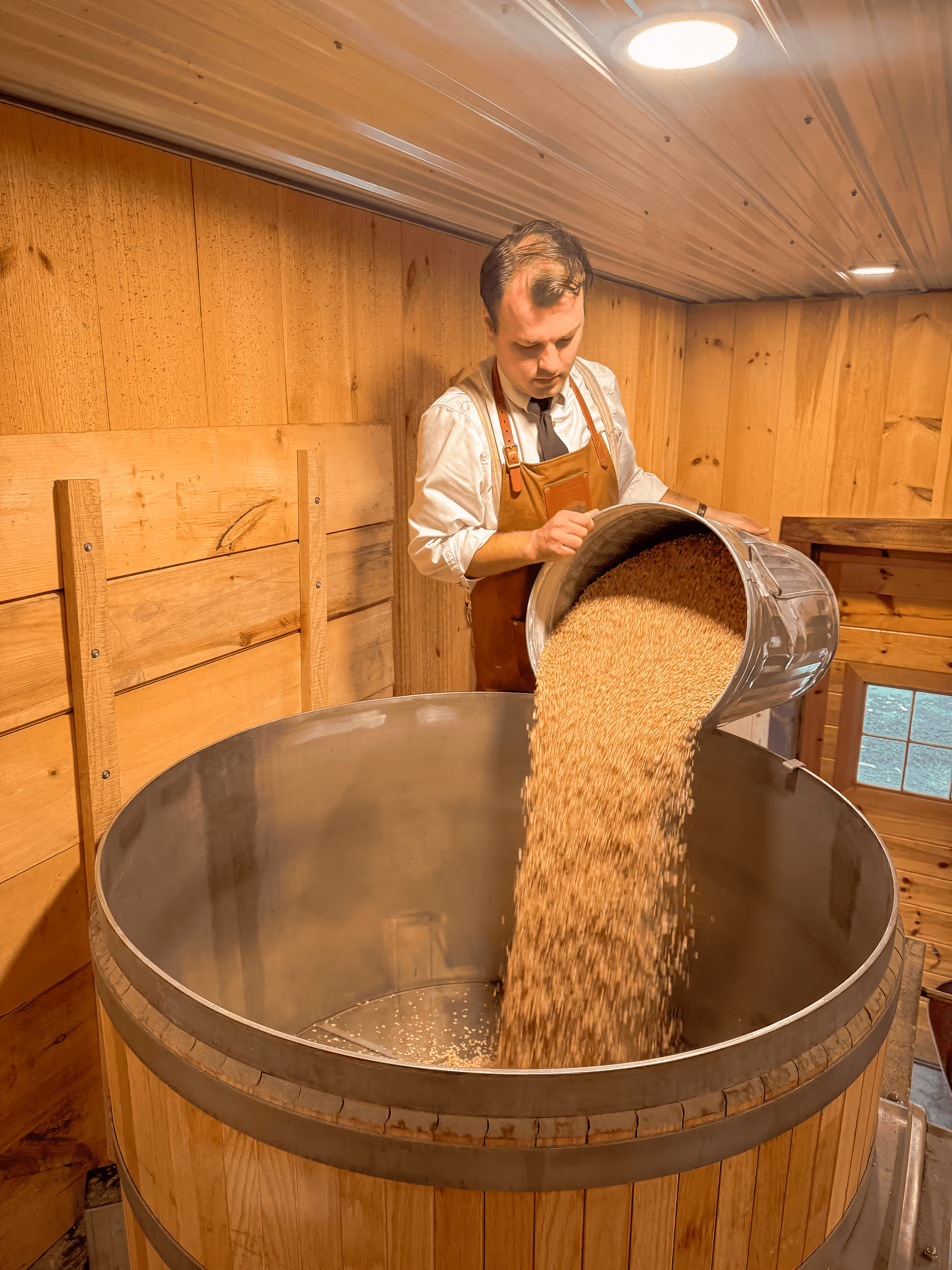 Craftsman pouring grain into large wooden barrel in rustic workshop