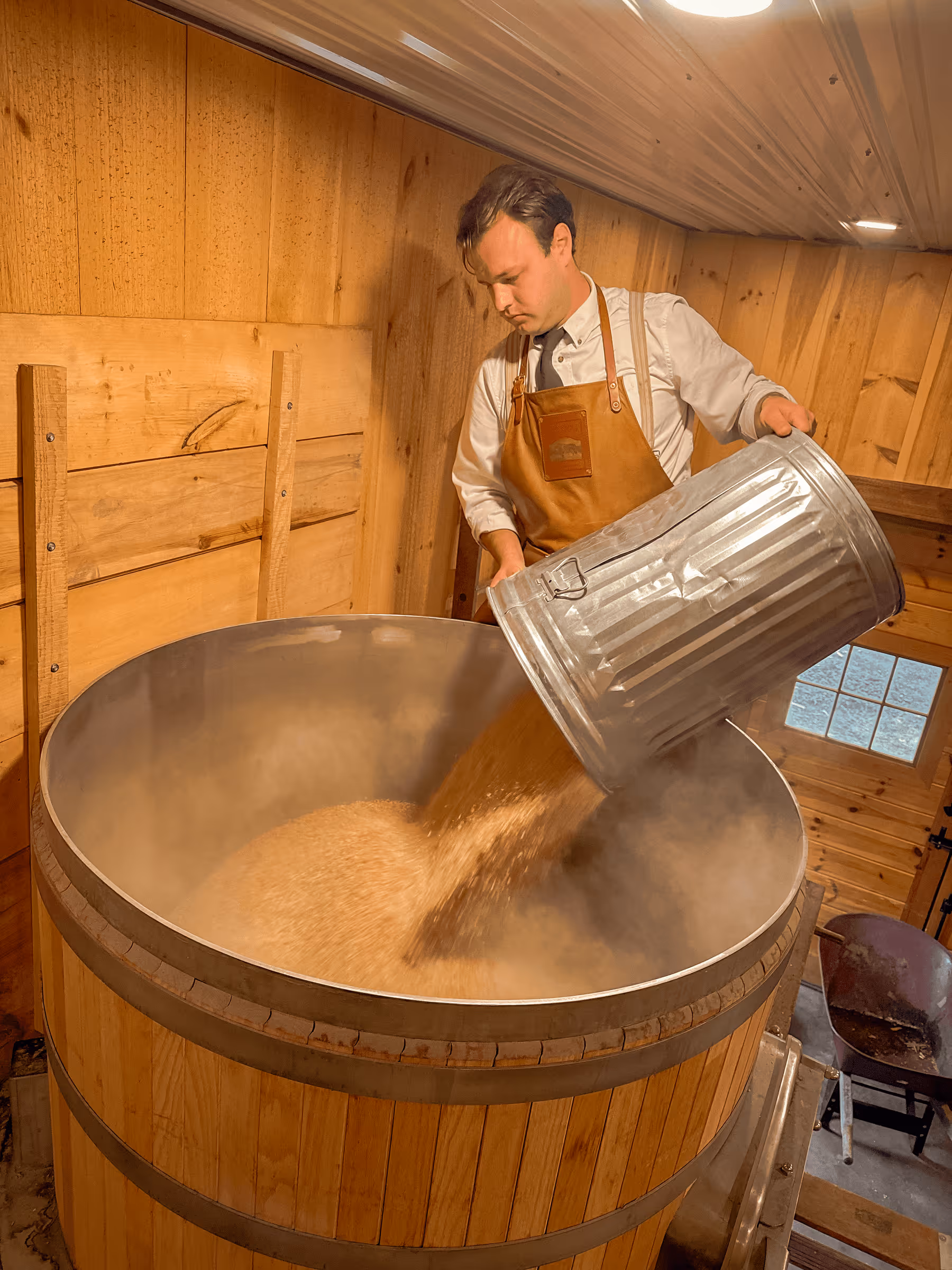 Brewer in apron pouring grains into large wooden brewing vessel