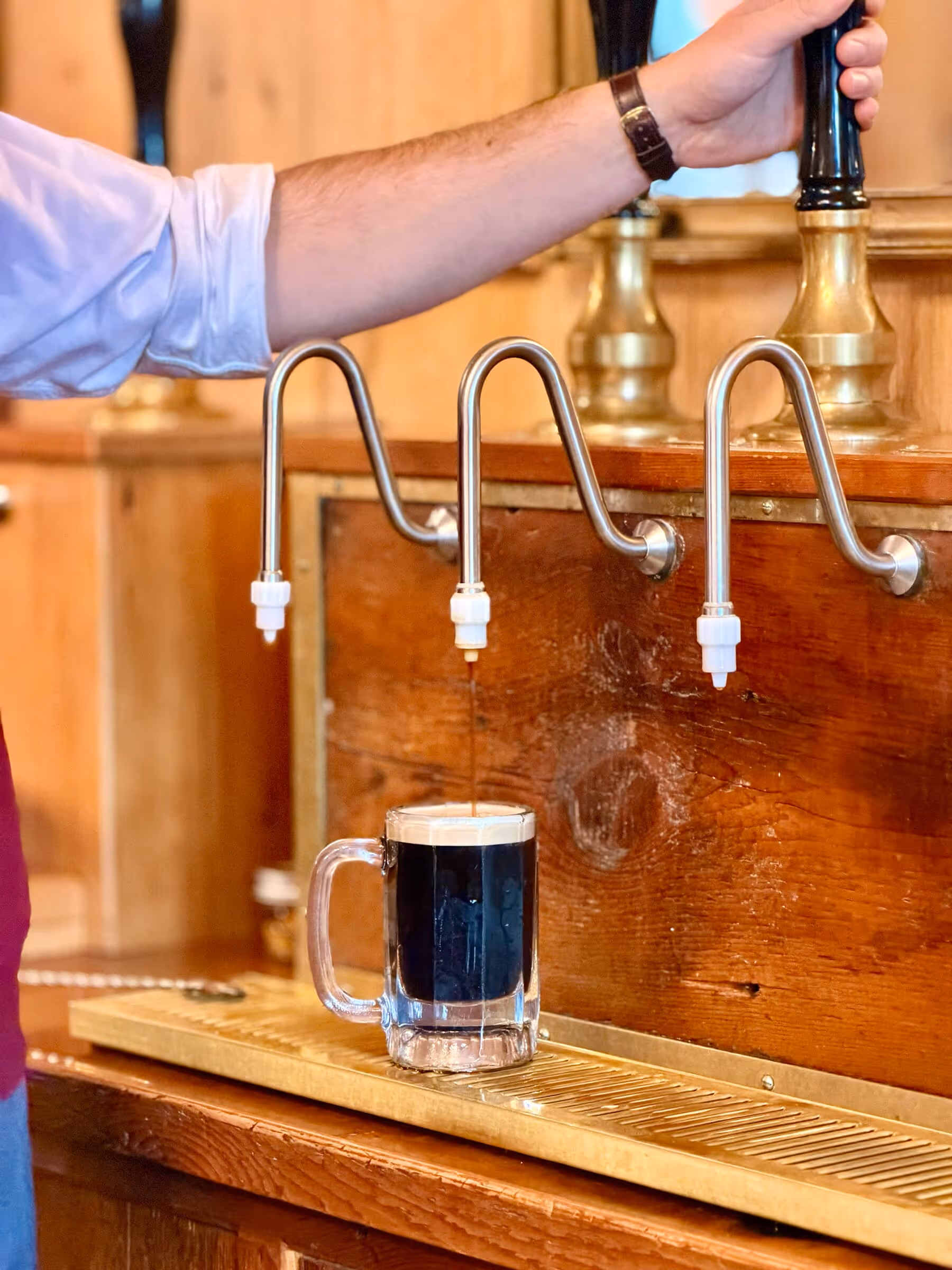 Person pouring a dark stout beer from a tap into a glass mug on a wooden bar.