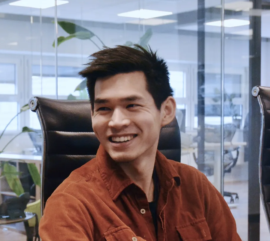 Smiling man with dark hair wearing a brown shirt sitting on a black office chair in a modern glass-walled workspace.
