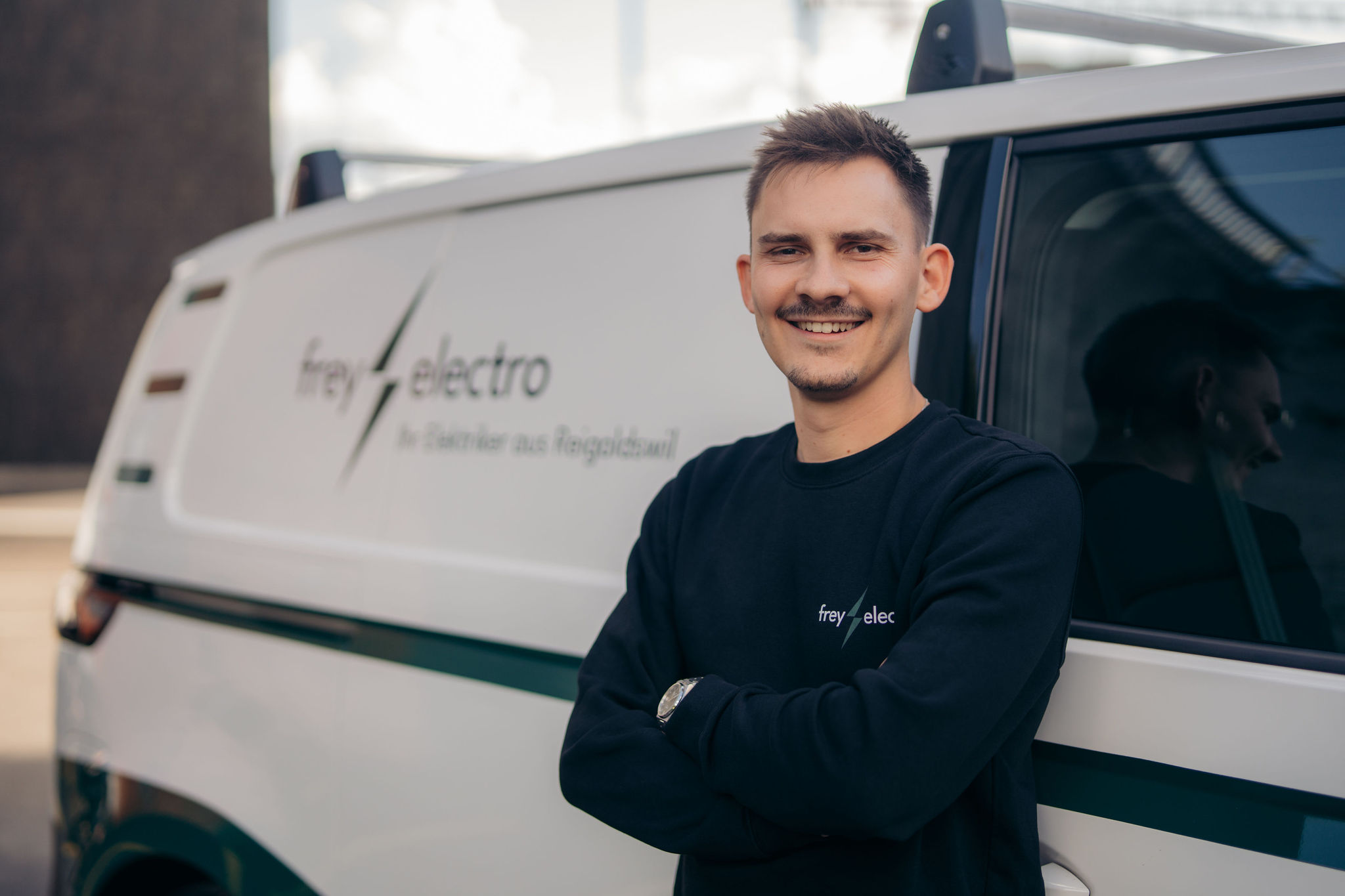 Junger Mann mit dunklem Pullover steht lächelnd mit verschränkten Armen vor einem weissen Fahrzeug mit dem Logo von Frey Electro.