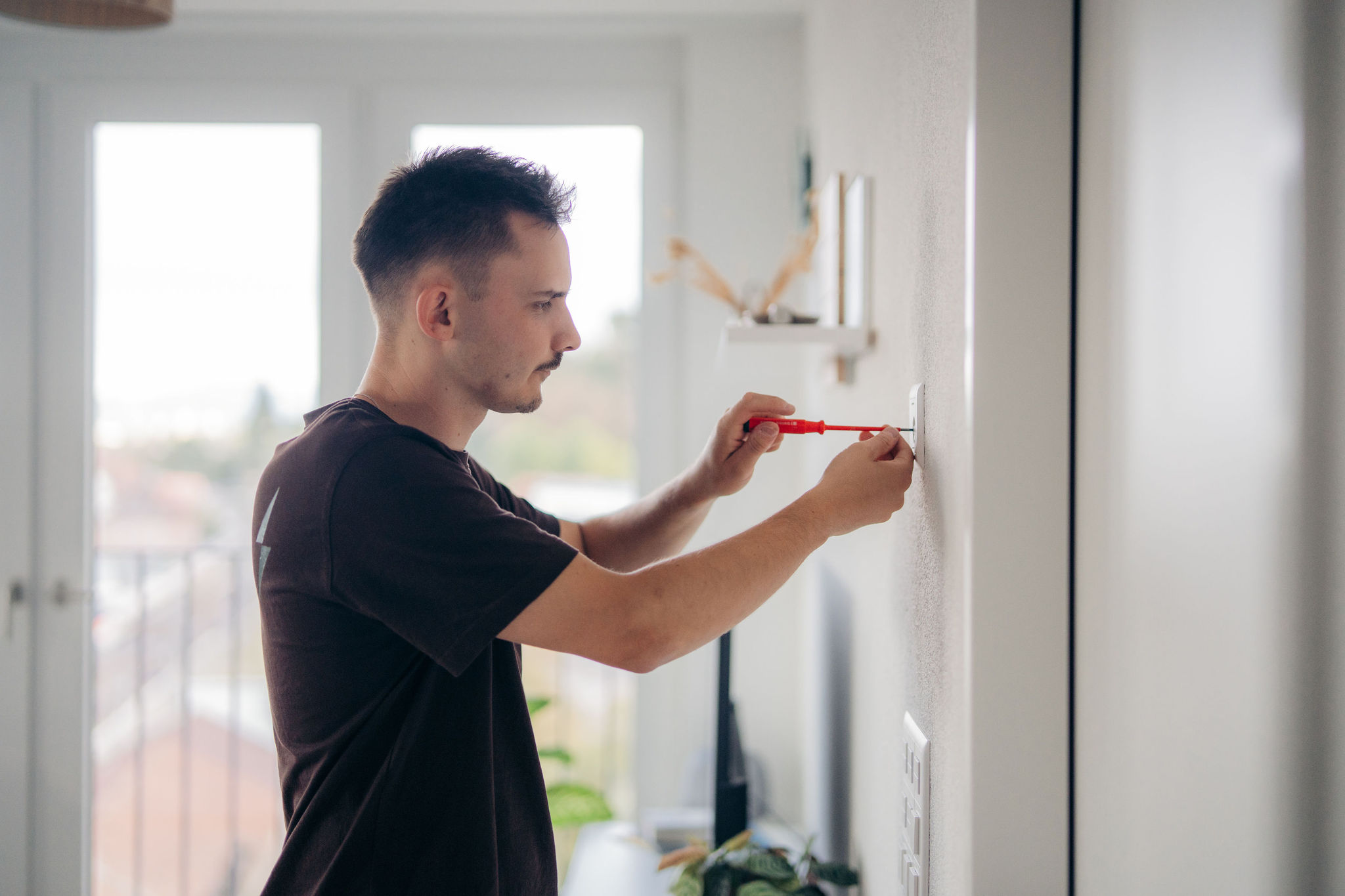 Junger Mann schraubt mit einem roten Schraubendreher eine Steckdosenabdeckung in einer hellen Wohnung an die Wand.