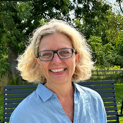 Lächelnde Frau mit blonden, kinnlangen Haaren, Brille und hellblauem Hemd vor grüner Baumkulisse.