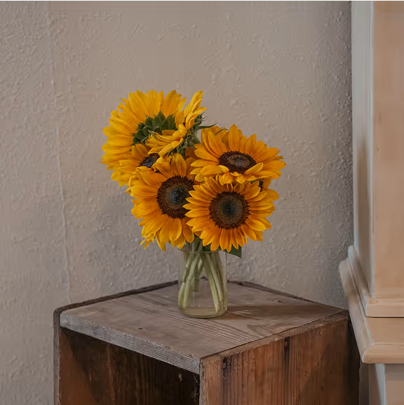 A bouquet of bright yellow sunflowers in a clear glass vase placed on a rustic wooden crate against a beige wall.
