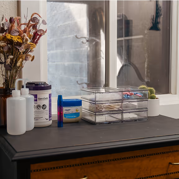 Black table with various skincare items including lotion bottles, a container of petroleum jelly, disinfecting wipes, a clear organizer with cotton pads and cotton swabs, a small potted cactus, and a vase with dried flowers in front of a window.