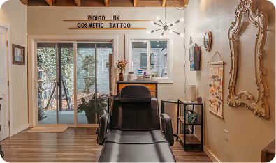 Interior of a cosmetic tattoo studio featuring a black tattoo chair, large decorative mirror, and a sign reading 'INDIGO INK COSMETIC TATTOO'.