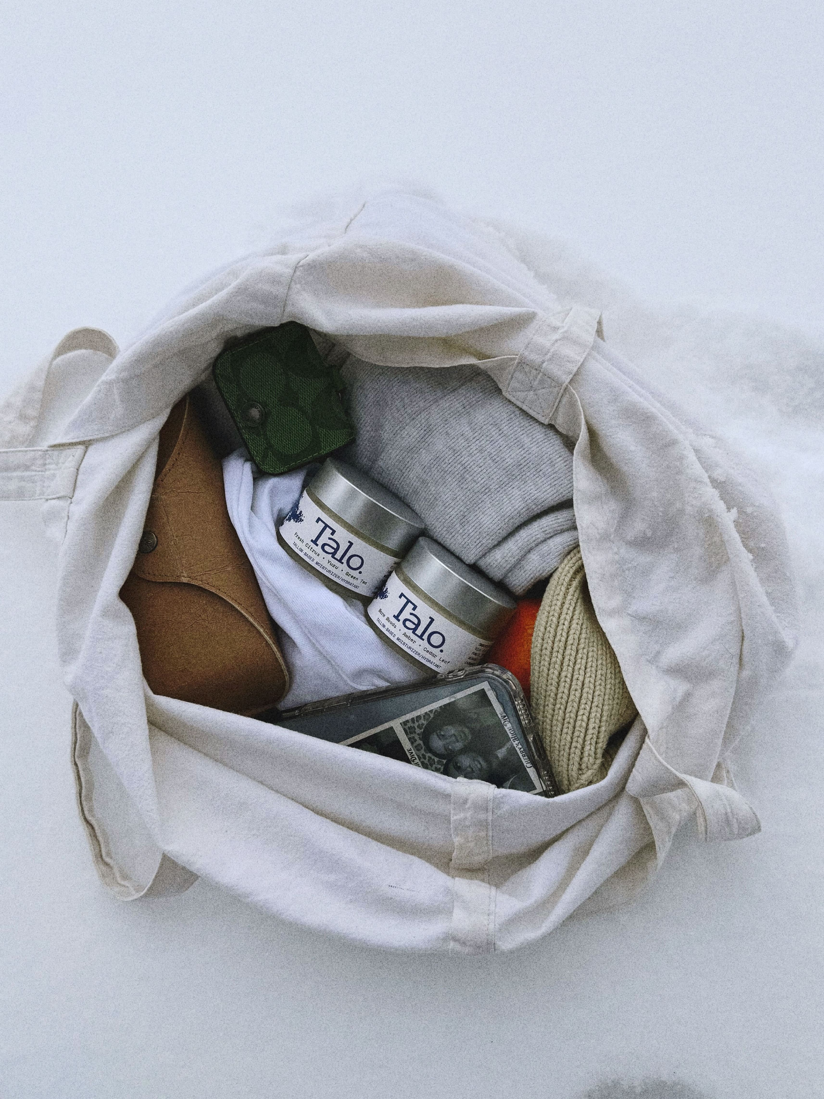 Open white tote bag on snow containing rolled clothes, two Talo skincare tins, a green pouch, and a cellphone.