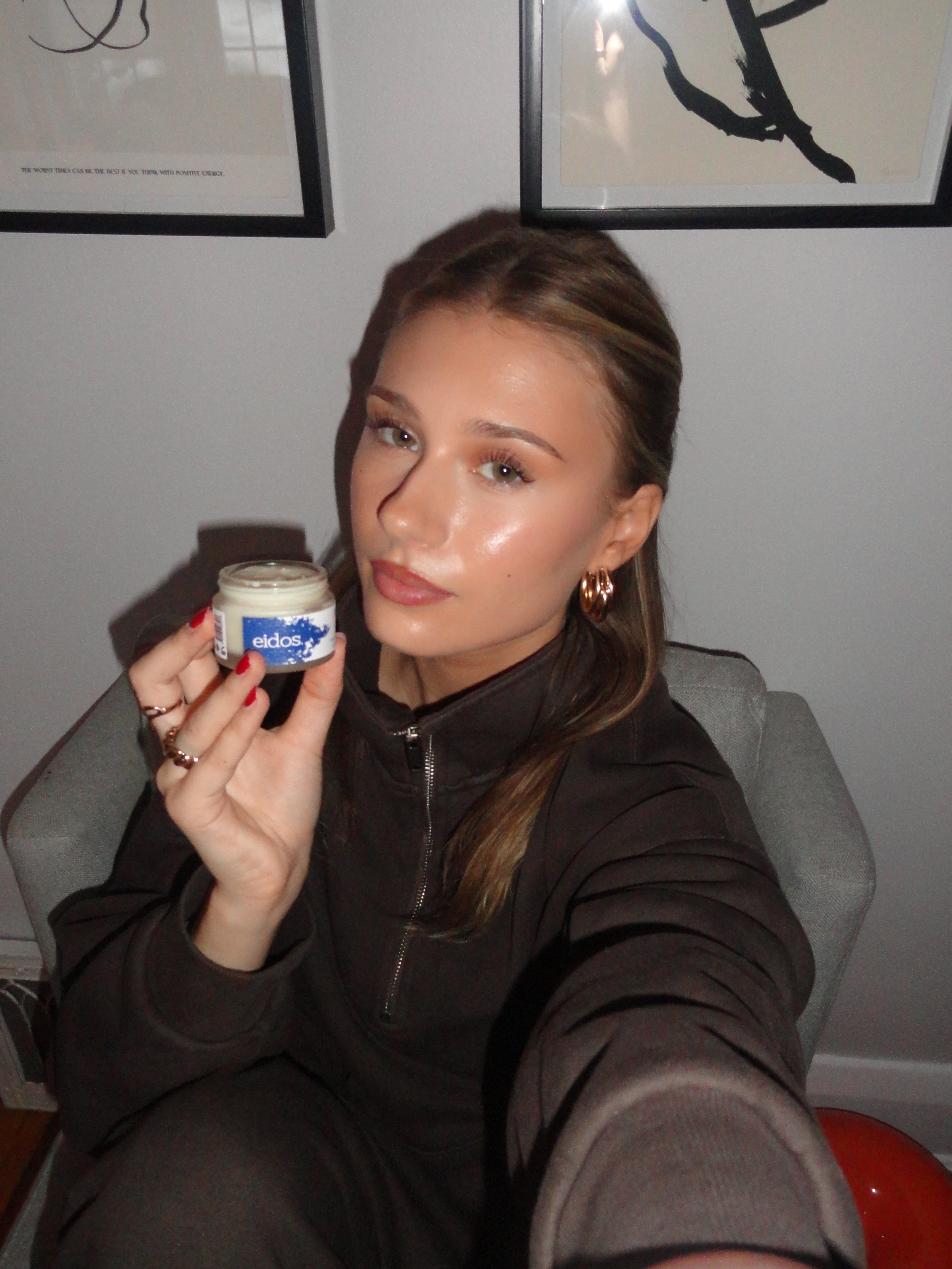 Young woman with light makeup and gold hoop earrings sitting in a chair, holding a small jar labeled 'eidos' toward the camera.