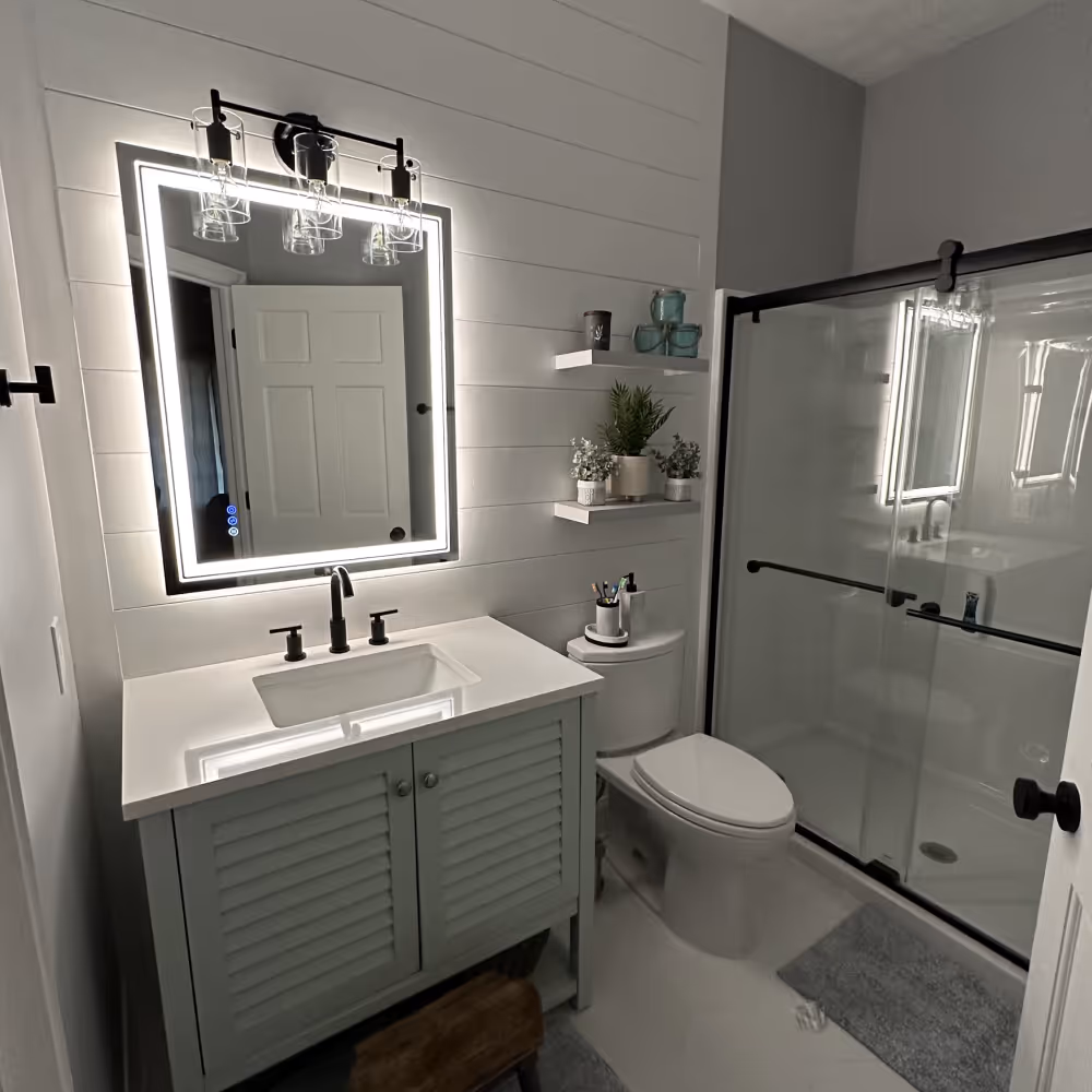 Modern bathroom with a lit rectangular mirror above a dark gray vanity, white sink, toilet, and glass shower with black frame done by J&J Renovation