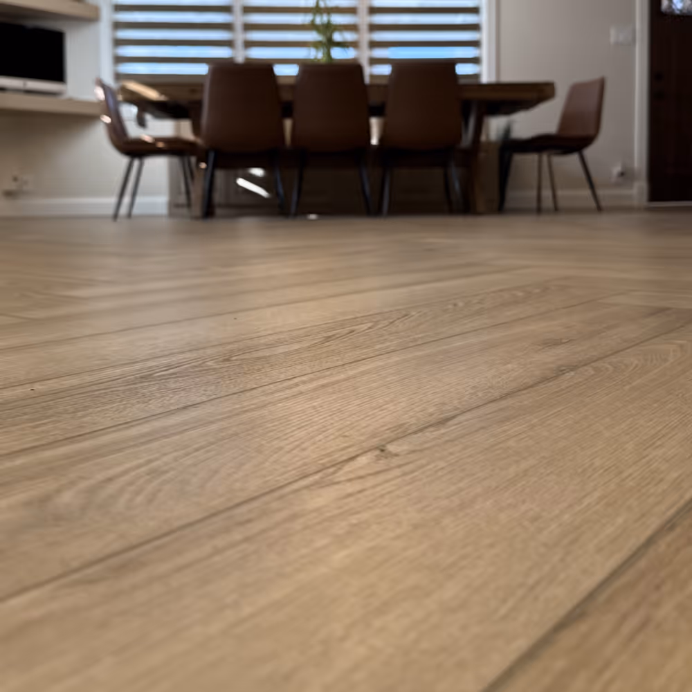 Close-up of light wood flooring in a dining room with a table and brown chairs in the background done by J&J Renovation
