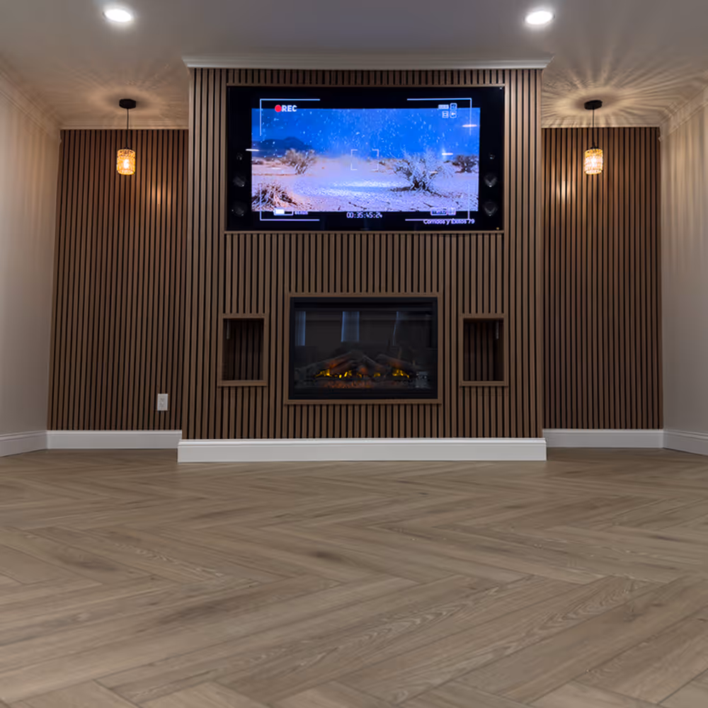 Modern living room wall with wood slat paneling, mounted flat-screen TV showing desert scene, electric fireplace below, and two pendant lights done by J&J Renovation