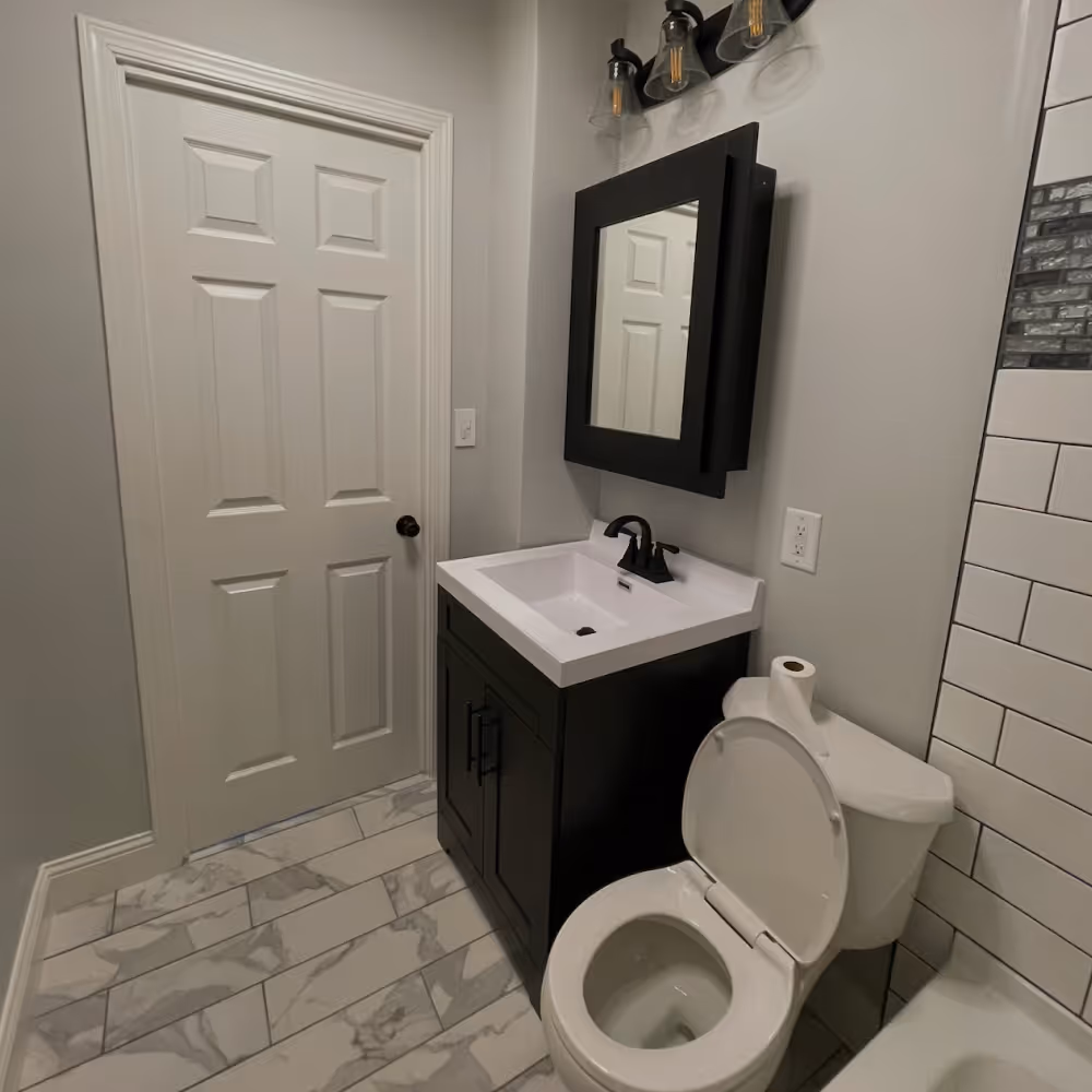 Modern bathroom with white toilet, black vanity with white sink, black framed mirror, and white door done by J&J Renovation.