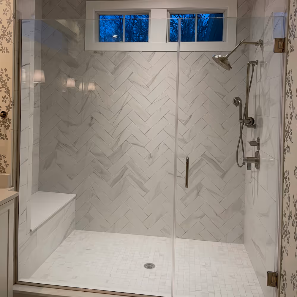 Modern walk-in shower with marble herringbone tile walls, built-in bench, and a stainless steel rain showerhead and handheld shower done by J&J Renovation
