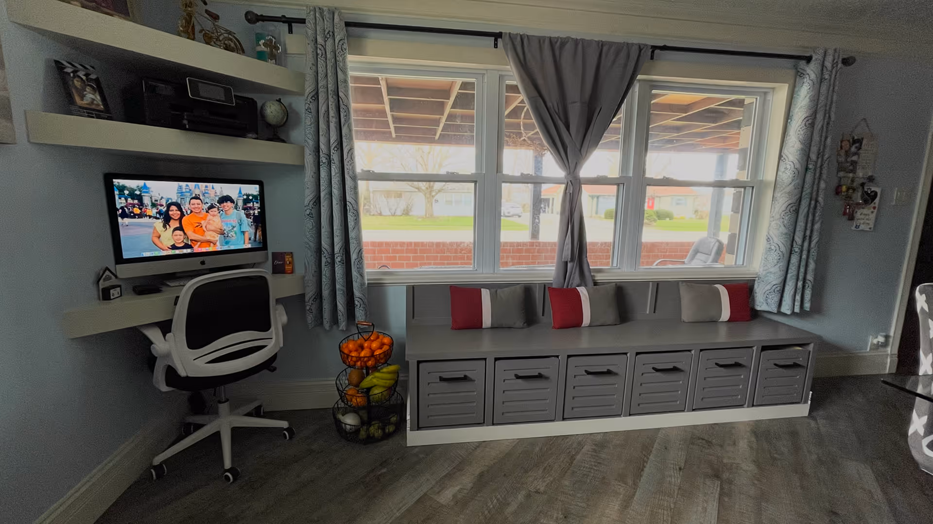 Modern home office corner with a computer desk and chair, family photo on the screen, gray built-in bench with storage drawers, decorative pillows, and a tiered fruit basket under the window done by J&J Renovation.