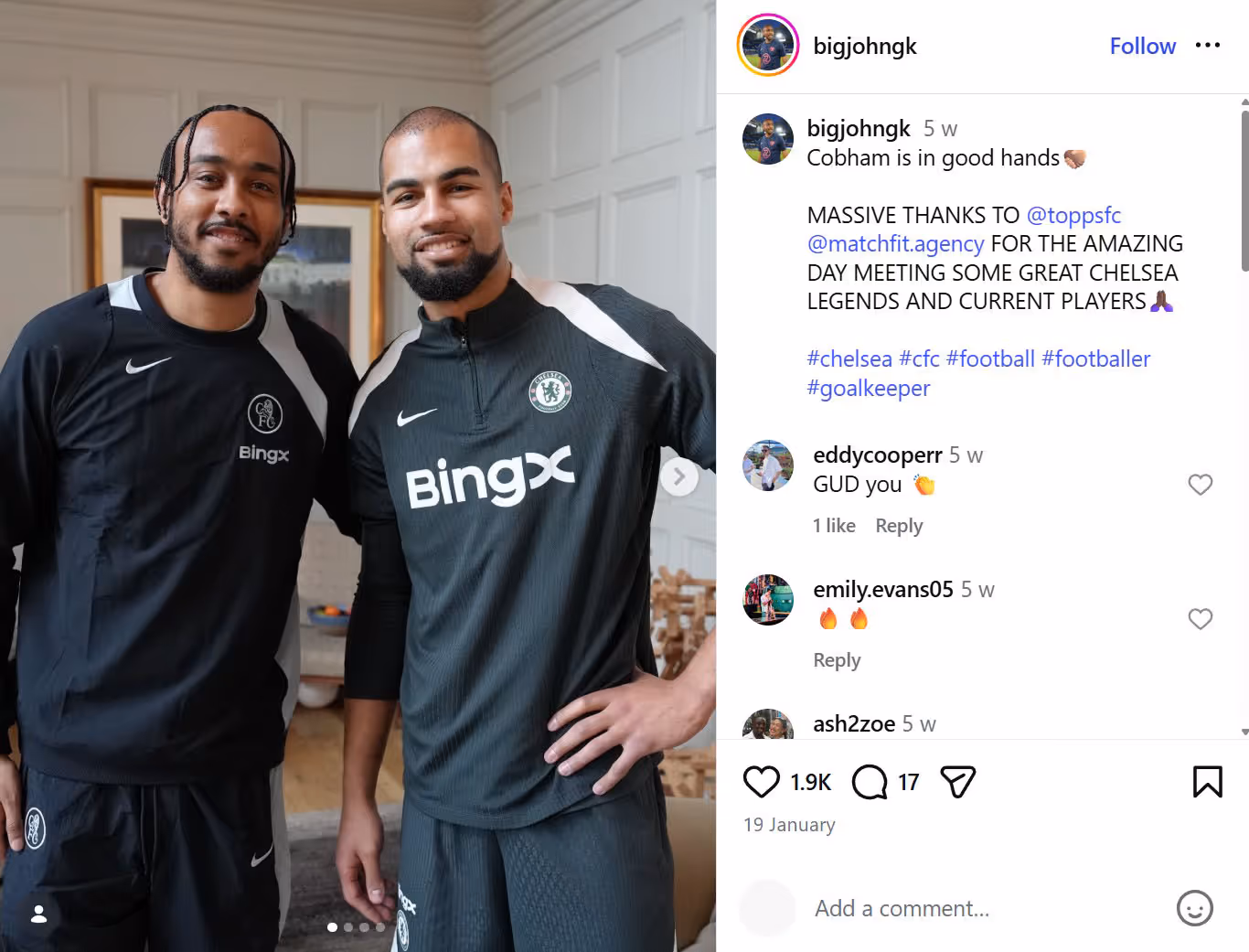 Two men in black Chelsea FC sportswear smiling and posing indoors against a white paneled wall.